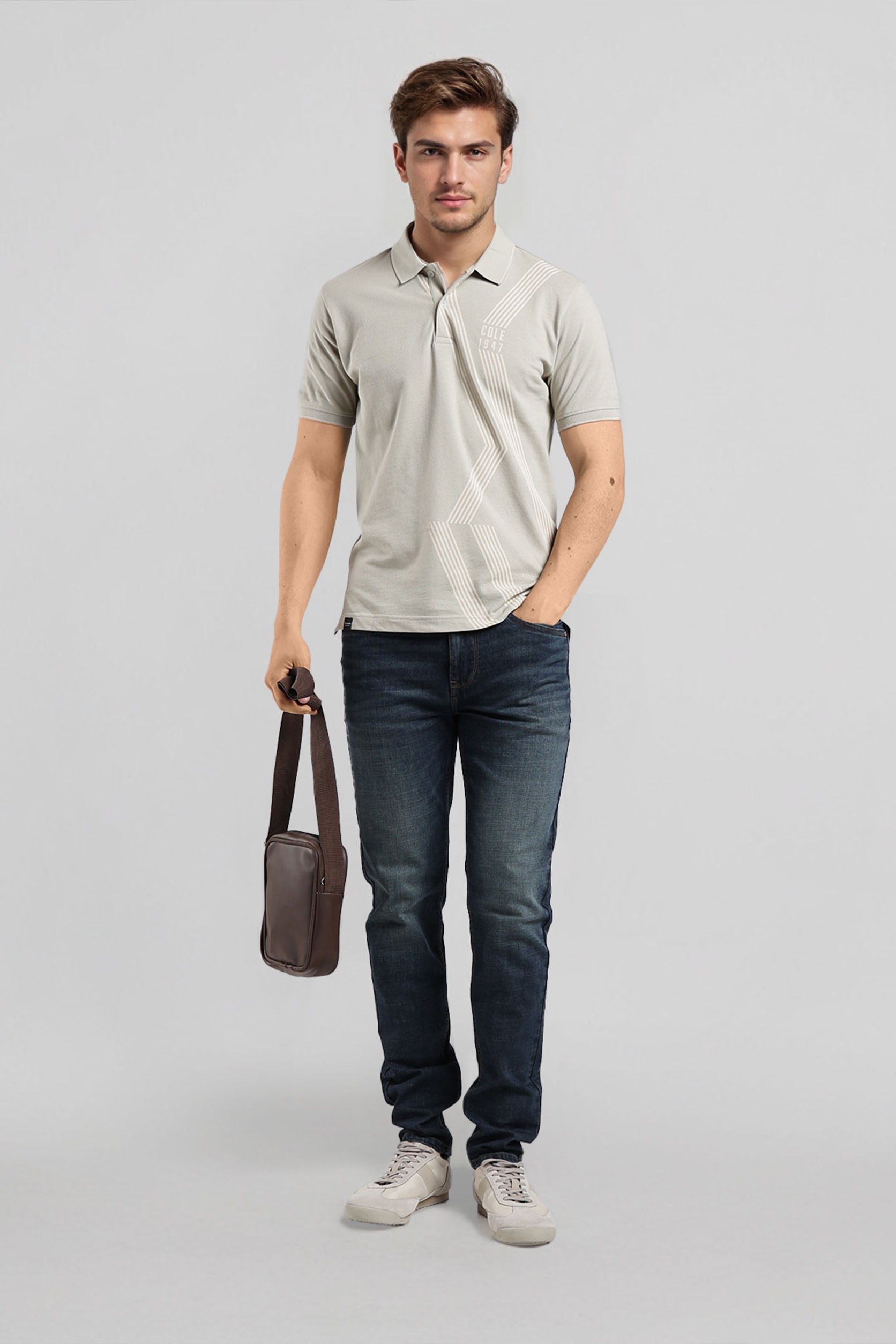 Man wearing a light gray polo shirt and dark blue jeans, holding a brown leather bag on a light gray background
