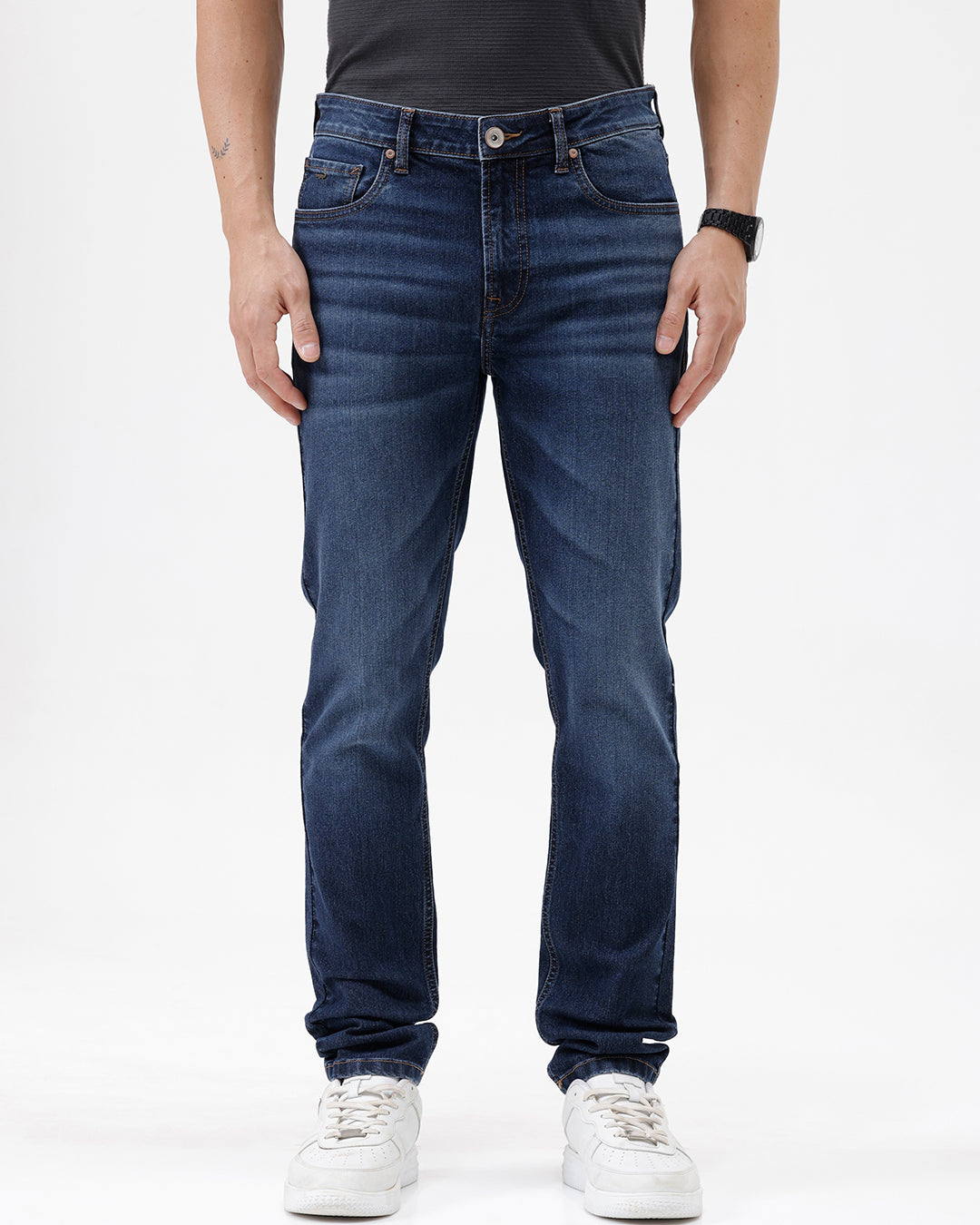 A person wearing dark blue slim-fit jeans with a faded wash, paired with a black t-shirt and white sneakers, shown from the waist down against a plain white studio background.