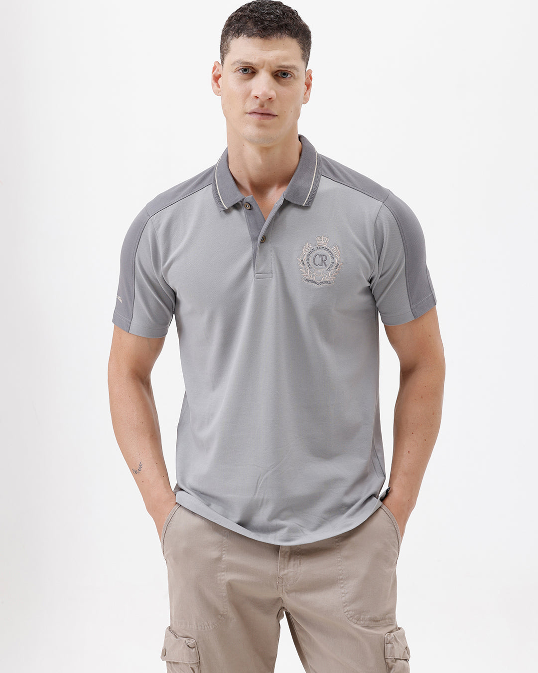 A man wearing a light gray short-sleeve polo shirt with darker gray shoulder panels and a small crest logo on the left chest, paired with khaki cargo pants, against a plain light gray studio background.