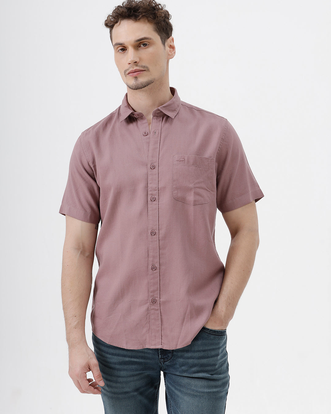 A man wearing a short-sleeve button-up shirt in dusty rose pink with dark blue jeans, standing against a plain white studio background.