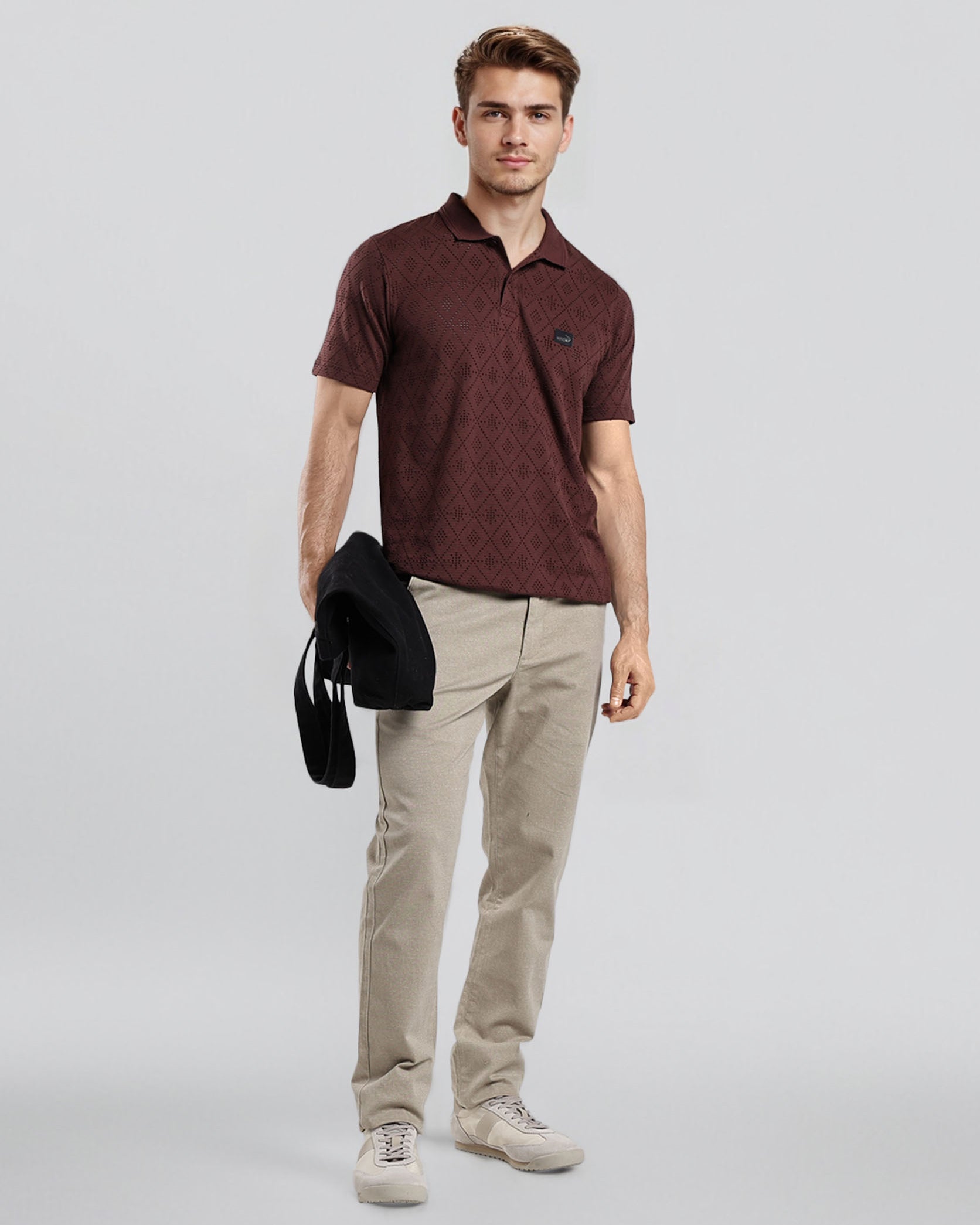 Man wearing a maroon polo shirt and beige pants holding a black jacket on a light gray background