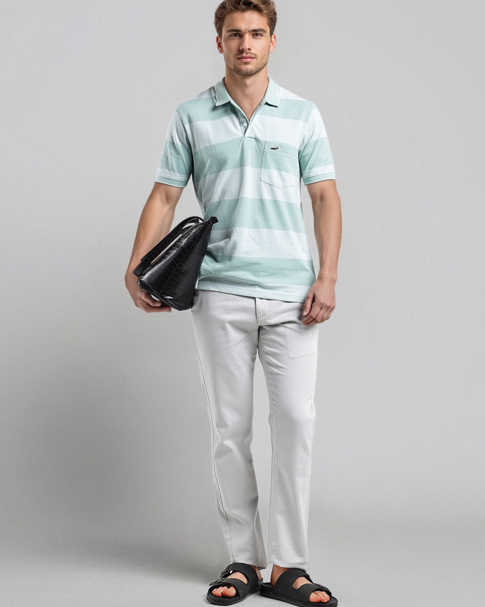 Man wearing a light blue striped polo shirt and white pants, holding a black bag on a gray background