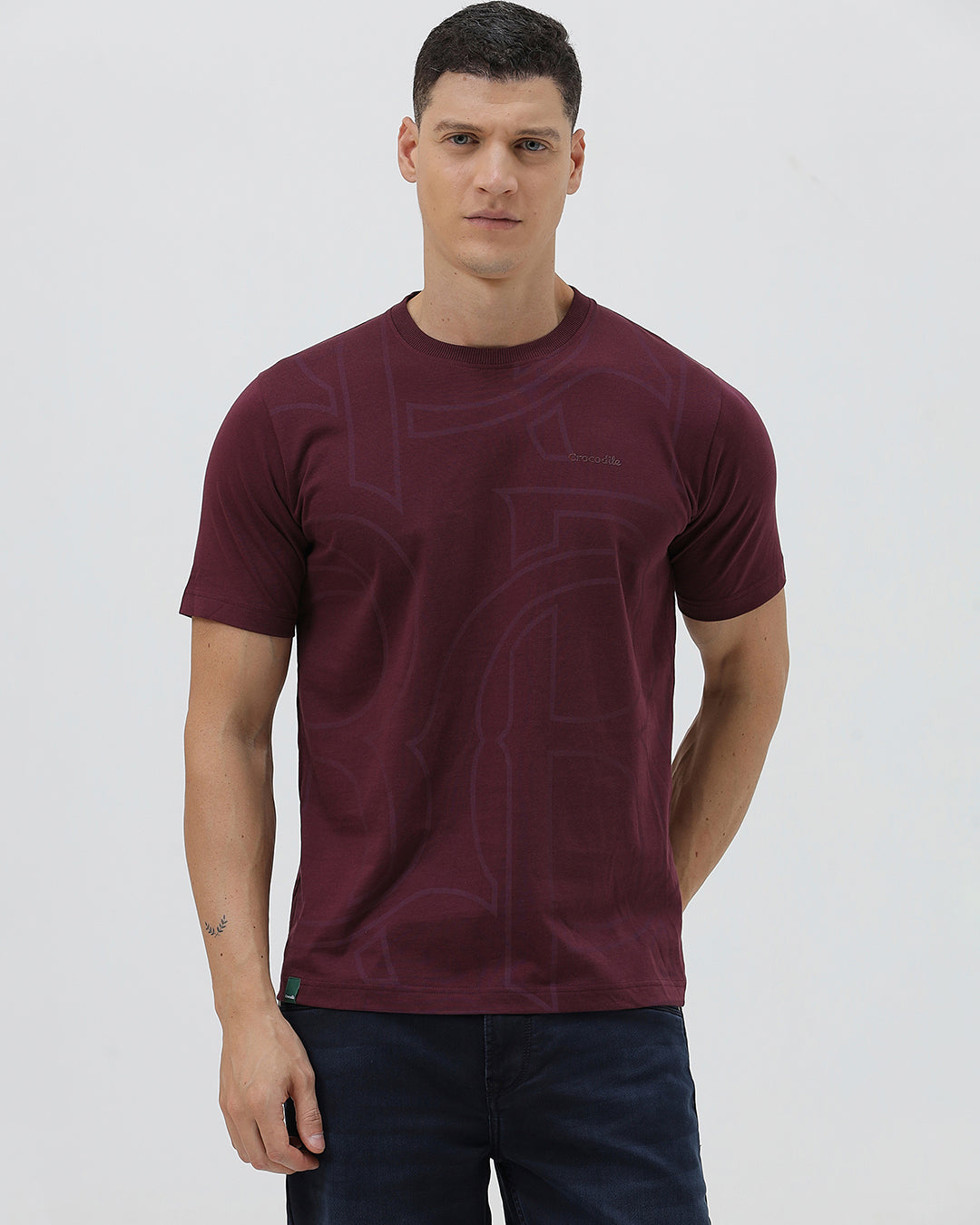 A man wearing a short-sleeve crewneck t-shirt in maroon with a subtle tonal graphic pattern, paired with dark blue jeans, against a light gray studio background.