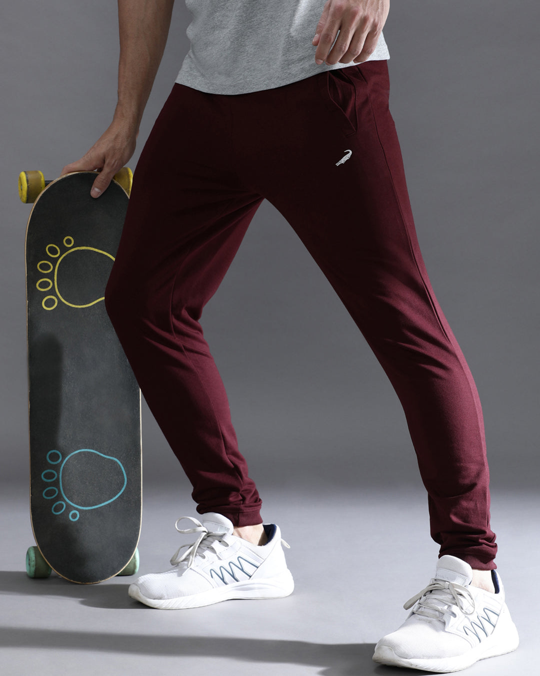 A person wearing wine red solid joggers with a comfort fit, standing next to a skateboard, wearing a grey top and white sneakers.
