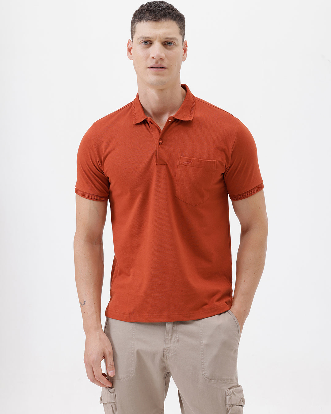 A man wearing a rust-orange short-sleeve polo shirt with a chest pocket, paired with beige cargo pants, standing against a plain light gray studio background.