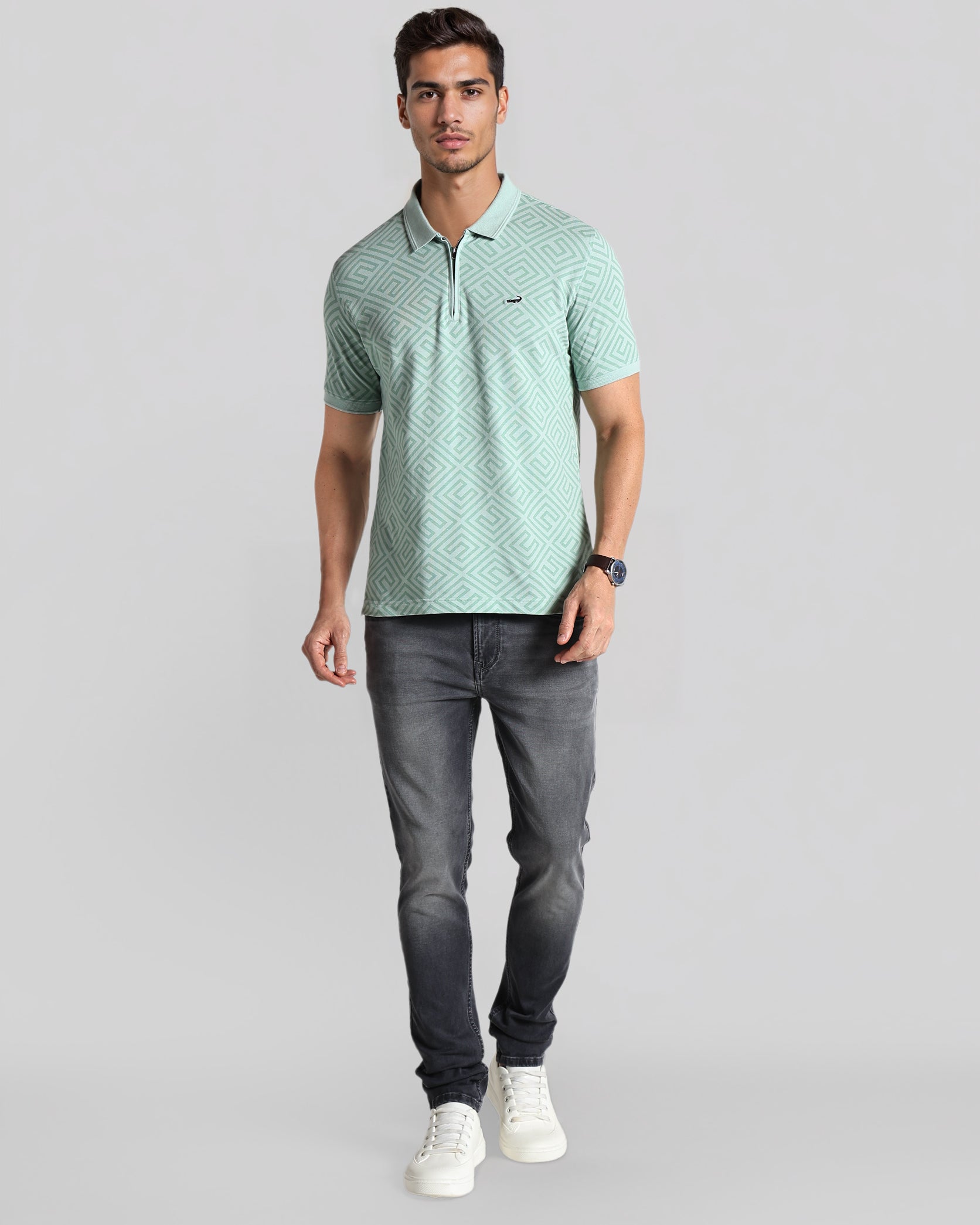 Man wearing a light green patterned polo shirt and dark gray jeans on a gray background