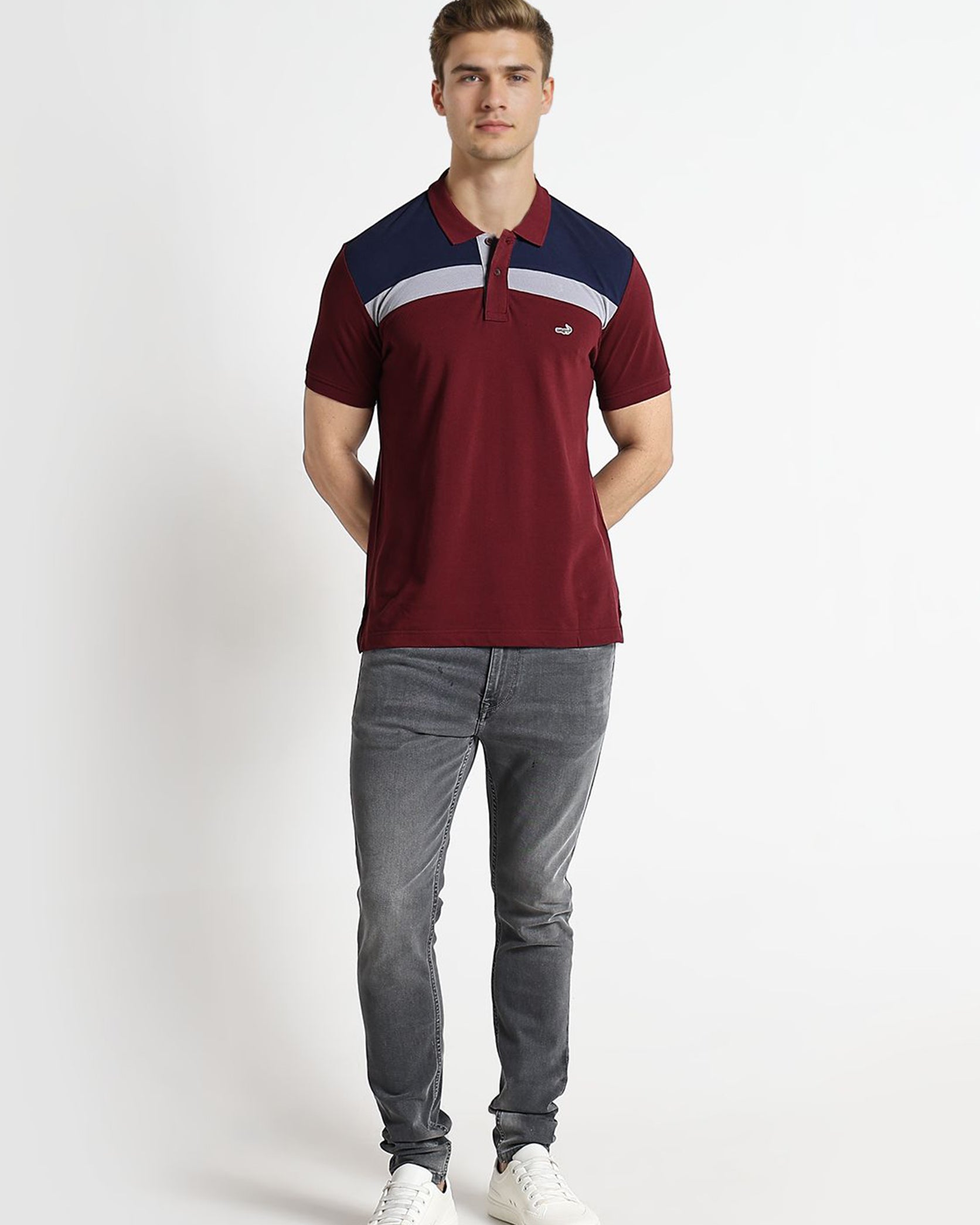 Man wearing a burgundy polo shirt with a blue and white stripe, gray jeans, and white sneakers on a white background