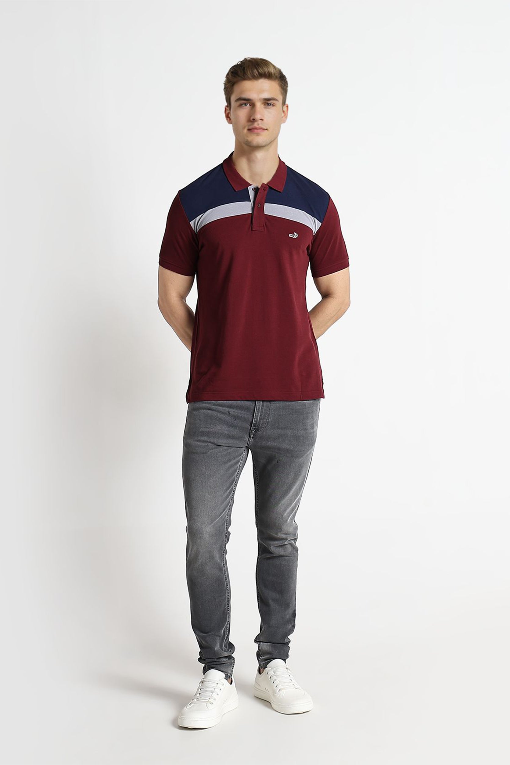 Man wearing a burgundy polo shirt with a blue and white stripe, gray jeans, and white sneakers on a white background