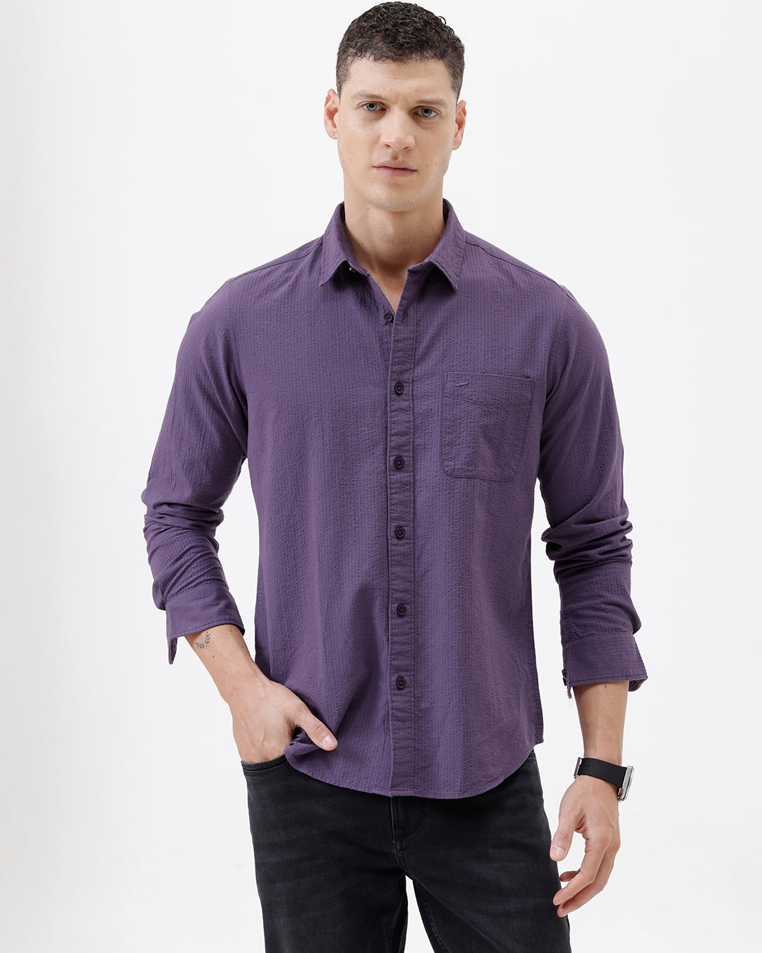 A man wearing a long-sleeve button-down shirt in a muted purple color, paired with black pants, standing against a plain white studio background.