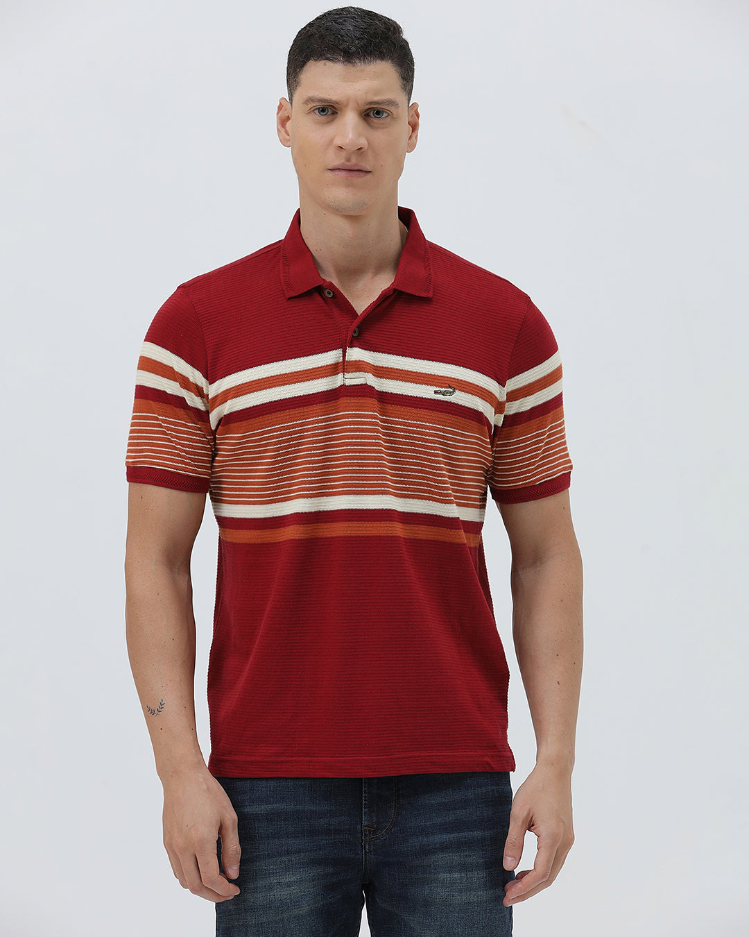 A man wearing a red short-sleeve polo shirt with horizontal cream and rust-orange stripes across the chest and sleeves, paired with dark blue jeans and black sunglasses, against a light gray studio background.