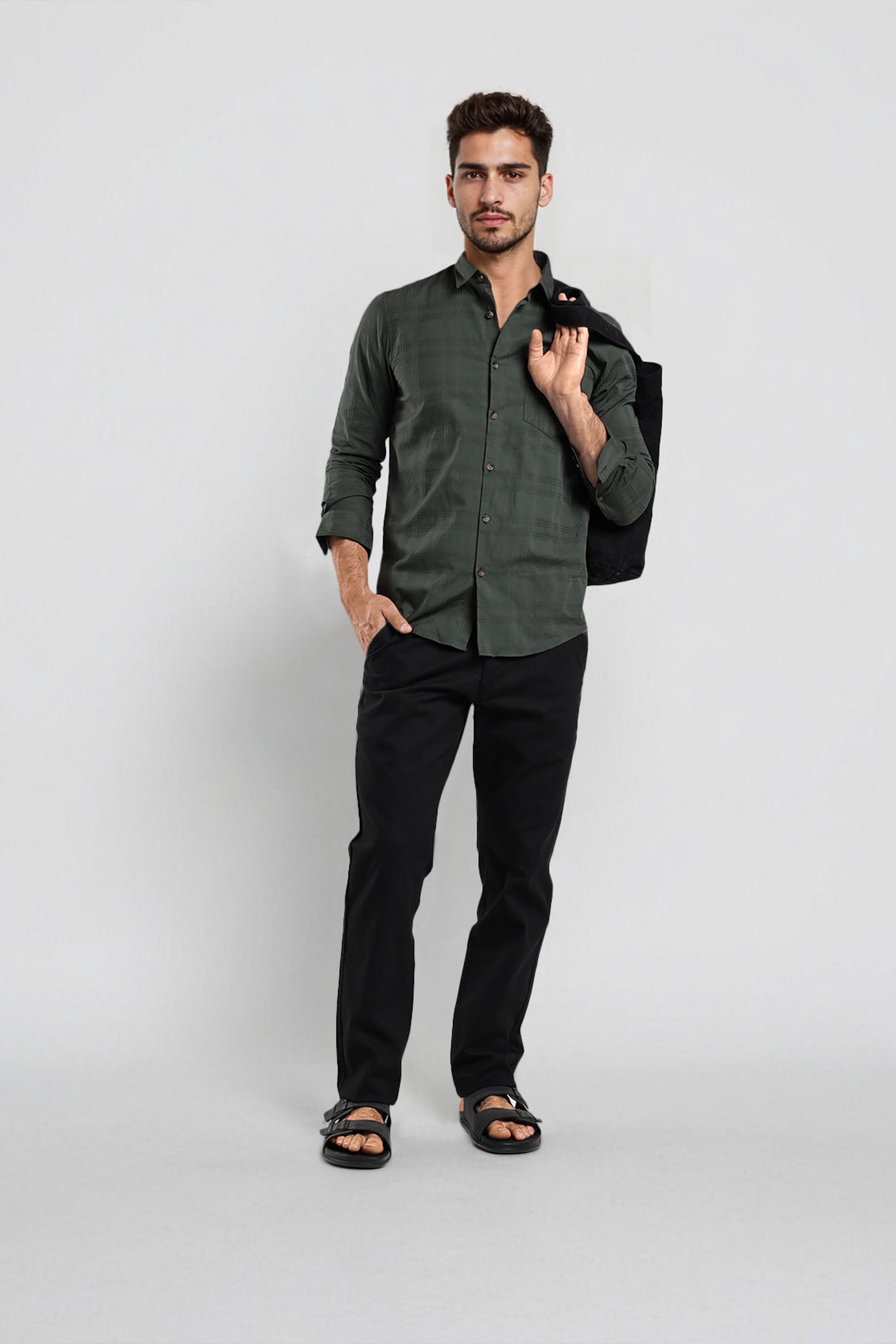 Man wearing a green shirt and black pants holding a black jacket against a white background