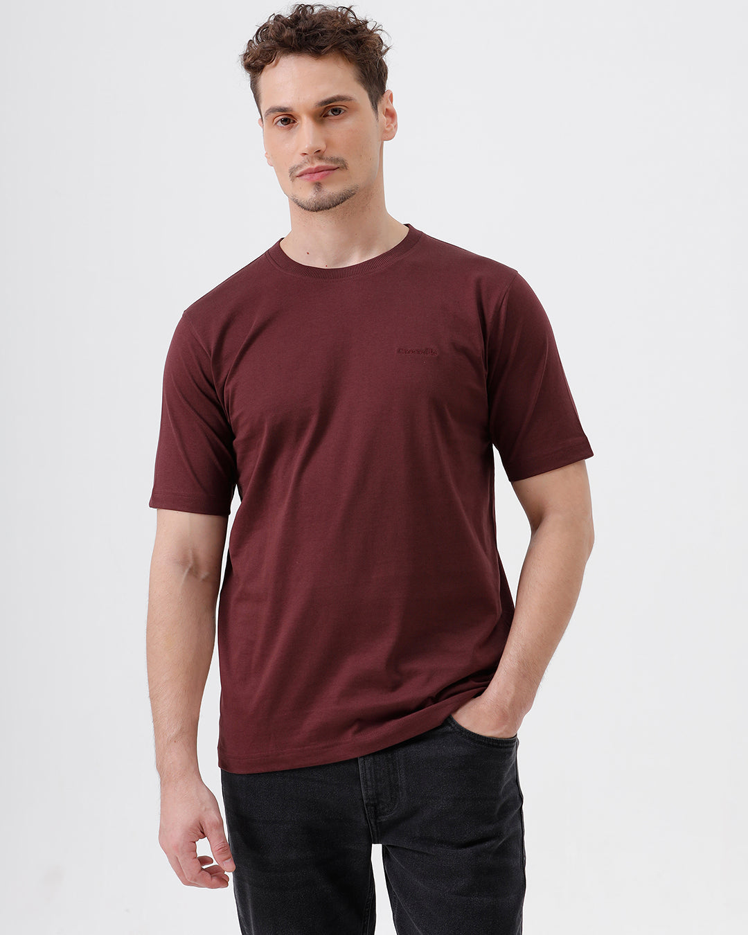 A man wearing a short-sleeve crewneck t-shirt in maroon, paired with dark jeans, standing against a plain light gray studio background.