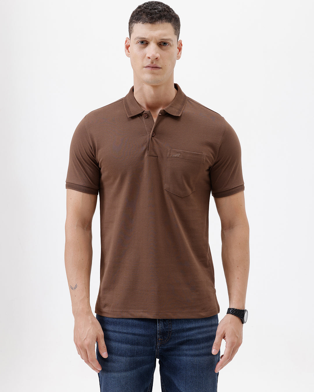 A man wearing a brown short-sleeve polo shirt with a chest pocket, paired with blue jeans, standing against a plain light gray background.