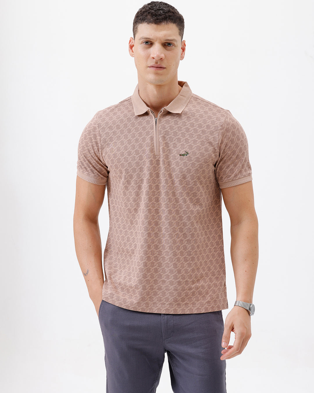 A man wearing a short-sleeve polo shirt with a muted brown and beige geometric pattern, paired with dark gray trousers, standing against a plain white studio background.