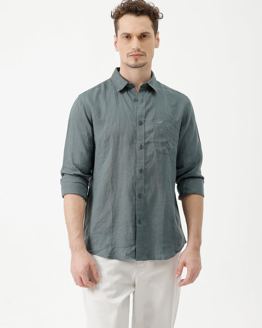 A man wearing a slate blue-green long-sleeve linen button-up shirt with white trousers, standing against a plain white studio background.