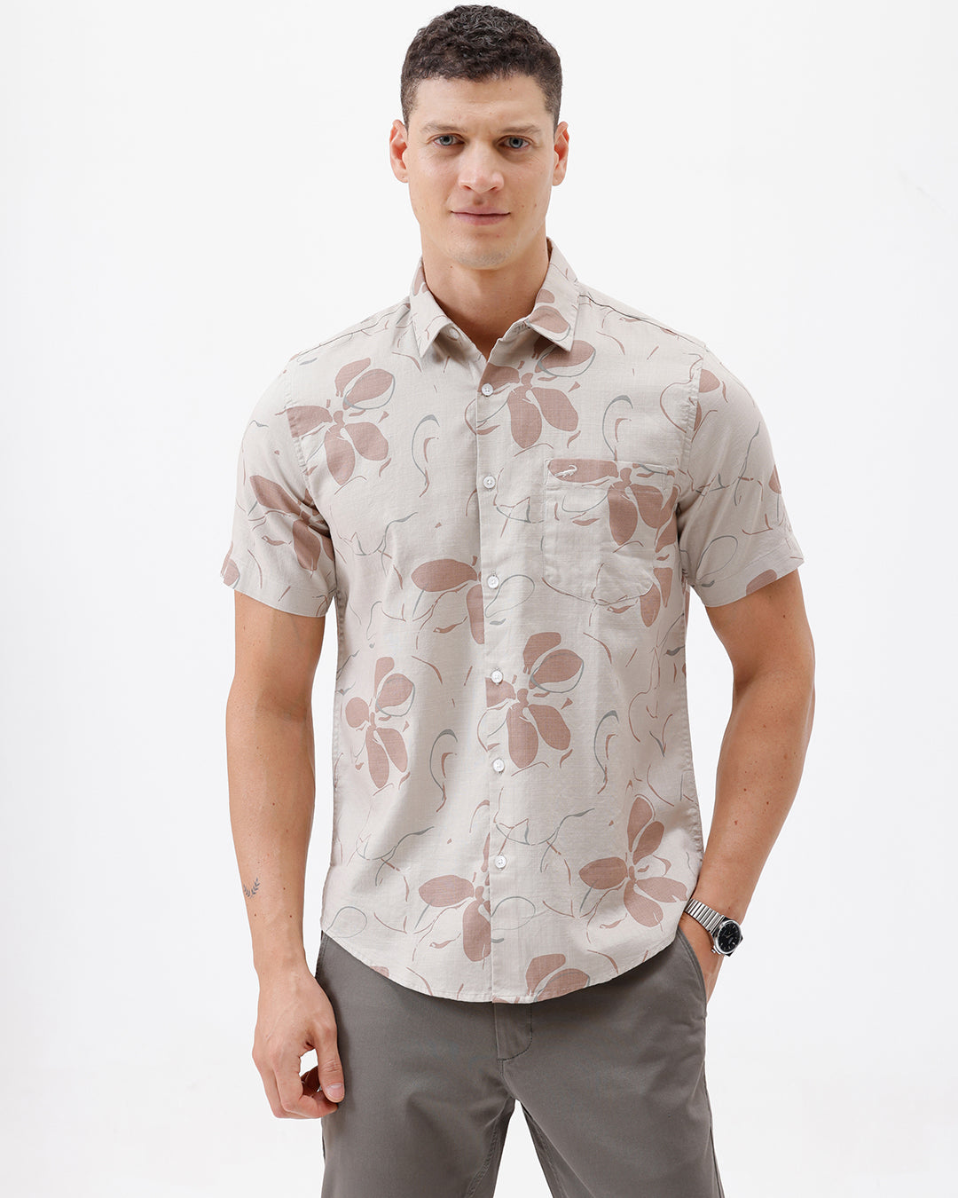 A man wearing a short-sleeve button-up shirt with a beige and brown floral pattern, paired with gray trousers, standing against a plain white background.