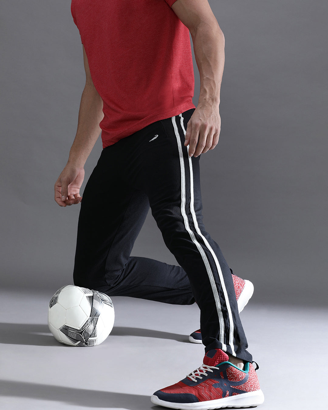 Person in red shirt and black pants with white stripes, kicking a soccer ball on a gray background