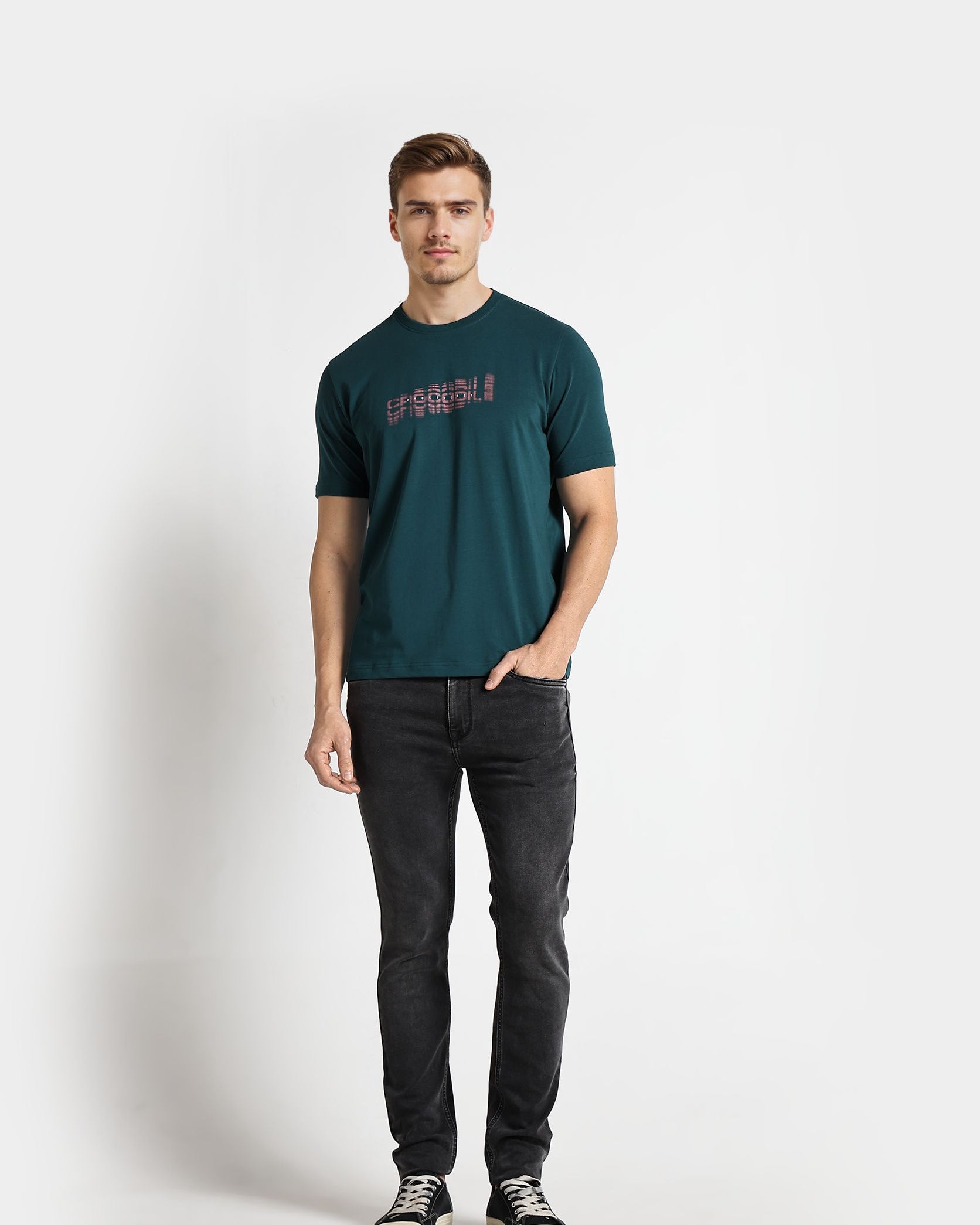 Man wearing a teal t-shirt with text and dark jeans on a white background