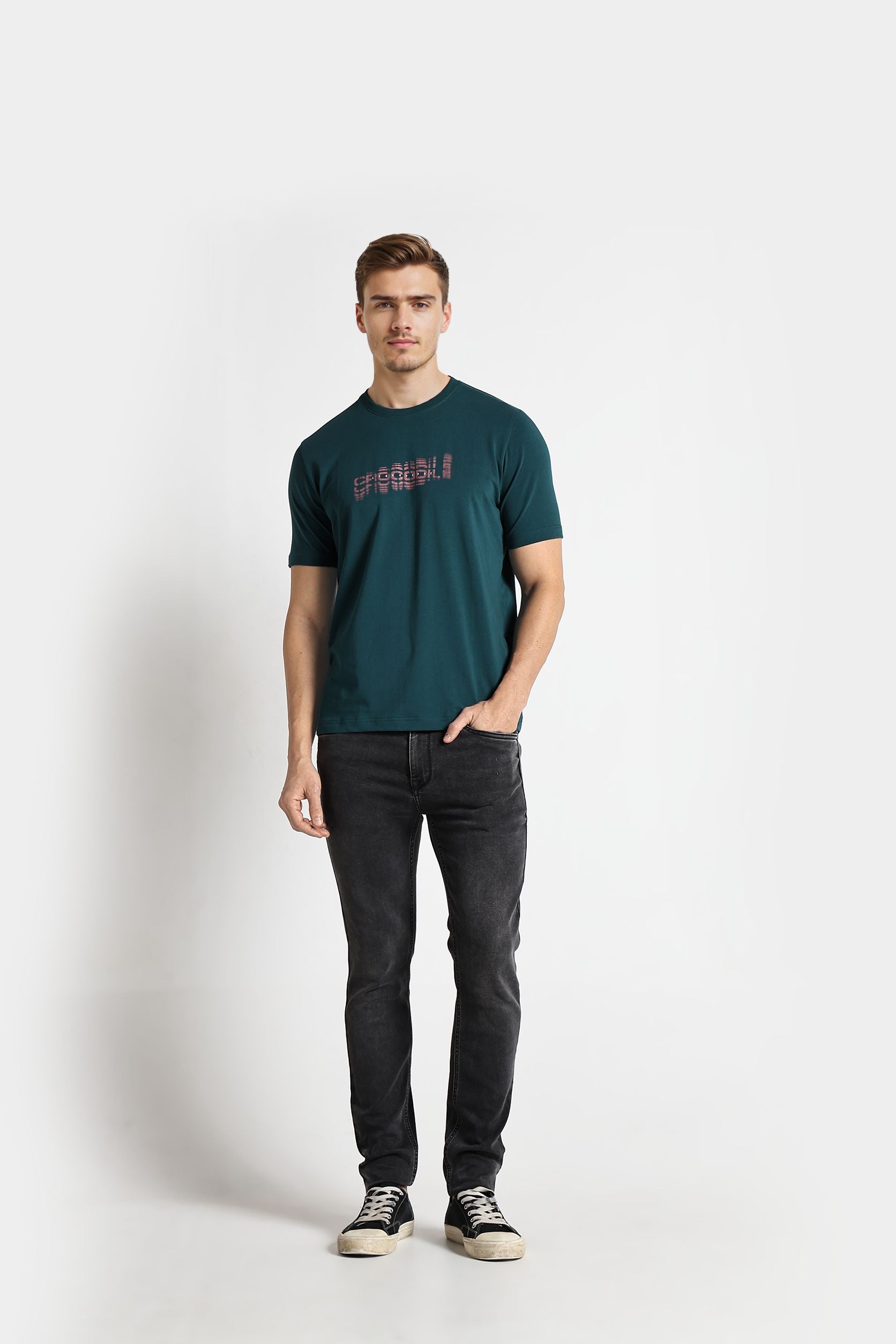 Man wearing a teal t-shirt with text and dark jeans on a white background
