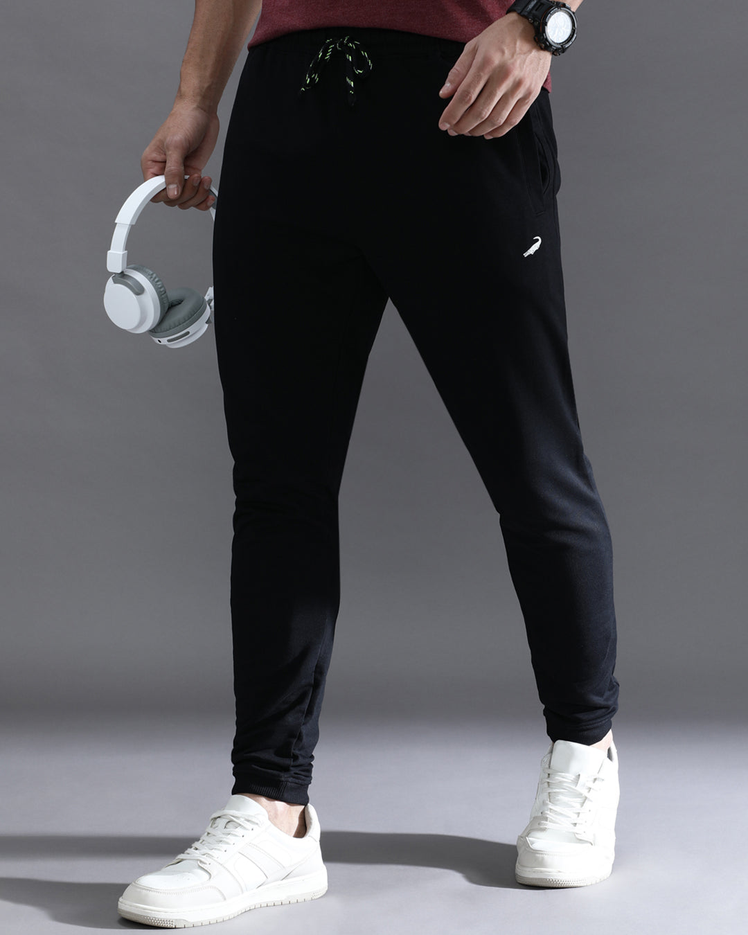 A person standing wearing solid black comfort fit joggers with a drawstring waist, paired with a red top and white sneakers. Accessories such as a watch and a keychain are visible.