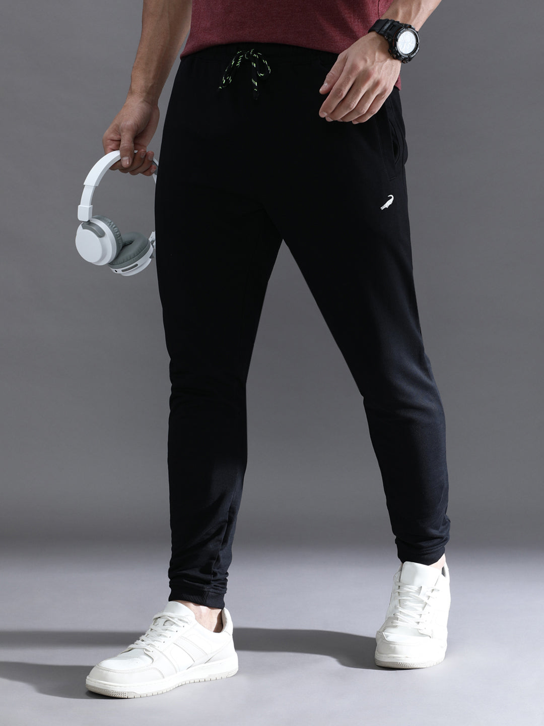 A person standing wearing solid black comfort fit joggers with a drawstring waist, paired with a red top and white sneakers. Accessories such as a watch and a keychain are visible.