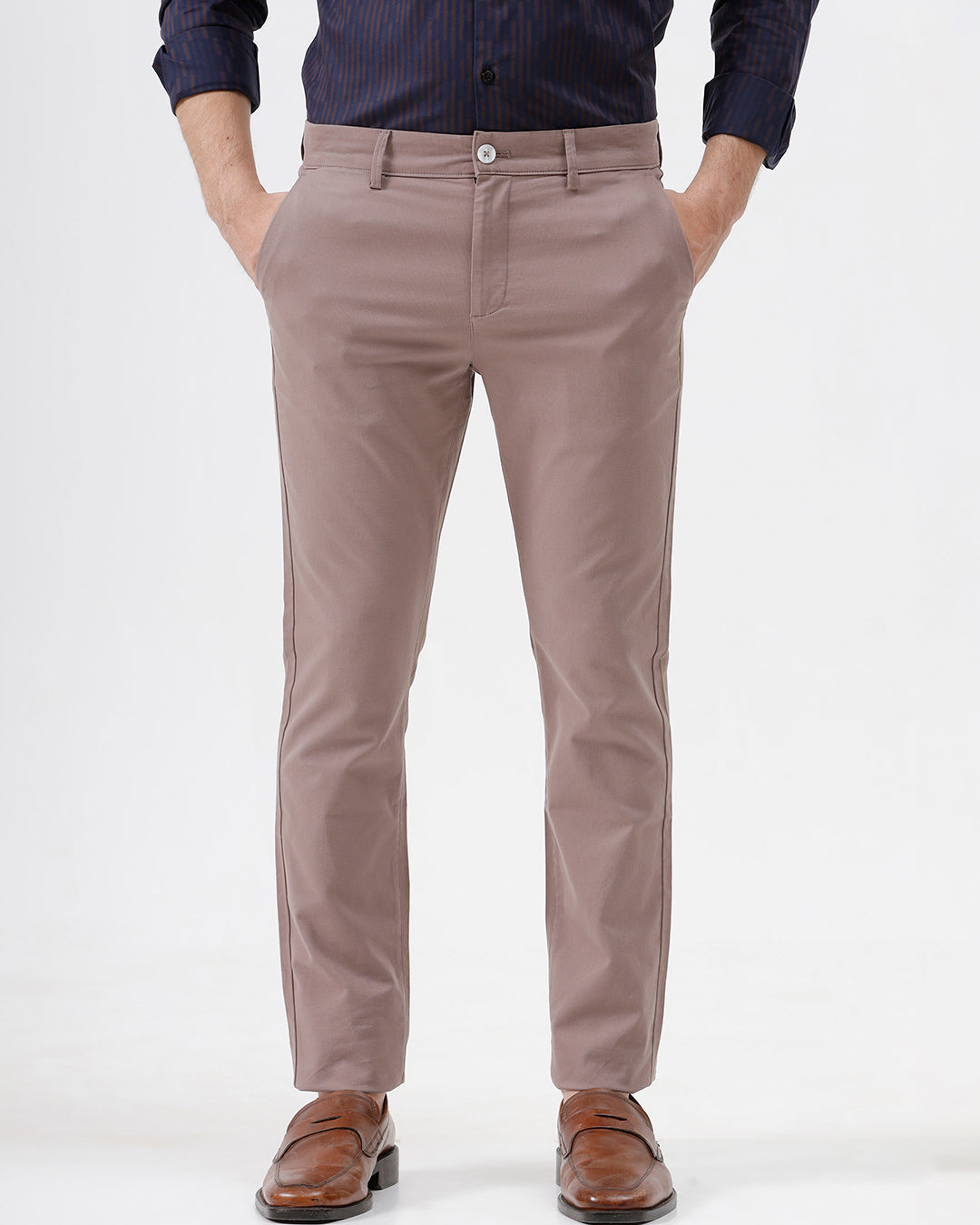 A person wearing slim-fit taupe trousers, a navy blue striped button-down shirt, and brown leather loafers, standing against a plain white studio background.