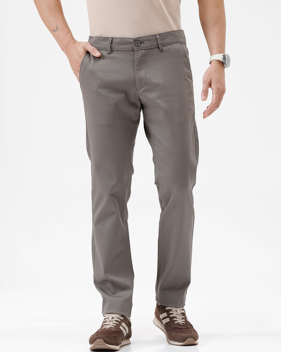 A man wearing slim-fit taupe gray chino pants, paired with a beige t-shirt and brown sneakers, shown from the waist down on a white studio background.