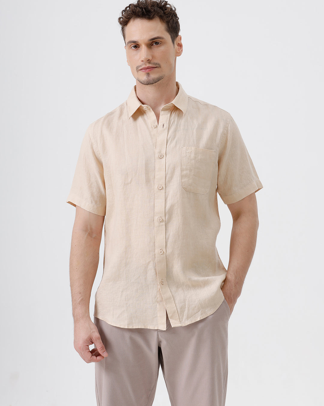A man wearing a light beige short-sleeve linen button-up shirt and taupe trousers, standing against a plain white studio background.