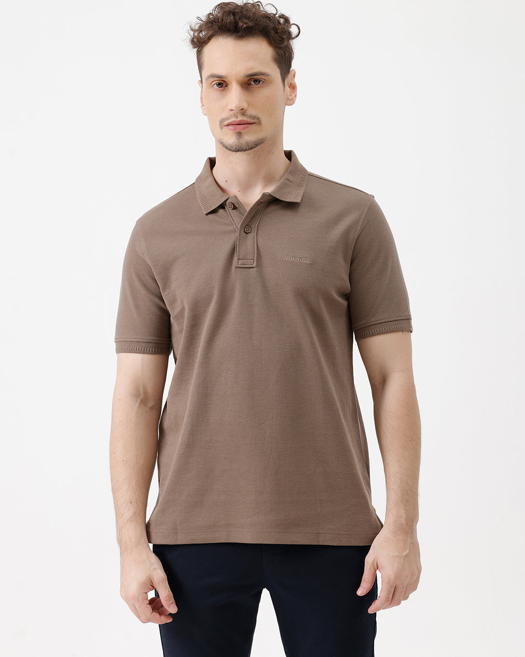 A man wearing a brown short-sleeve polo shirt with dark navy pants, standing against a plain light gray studio background.