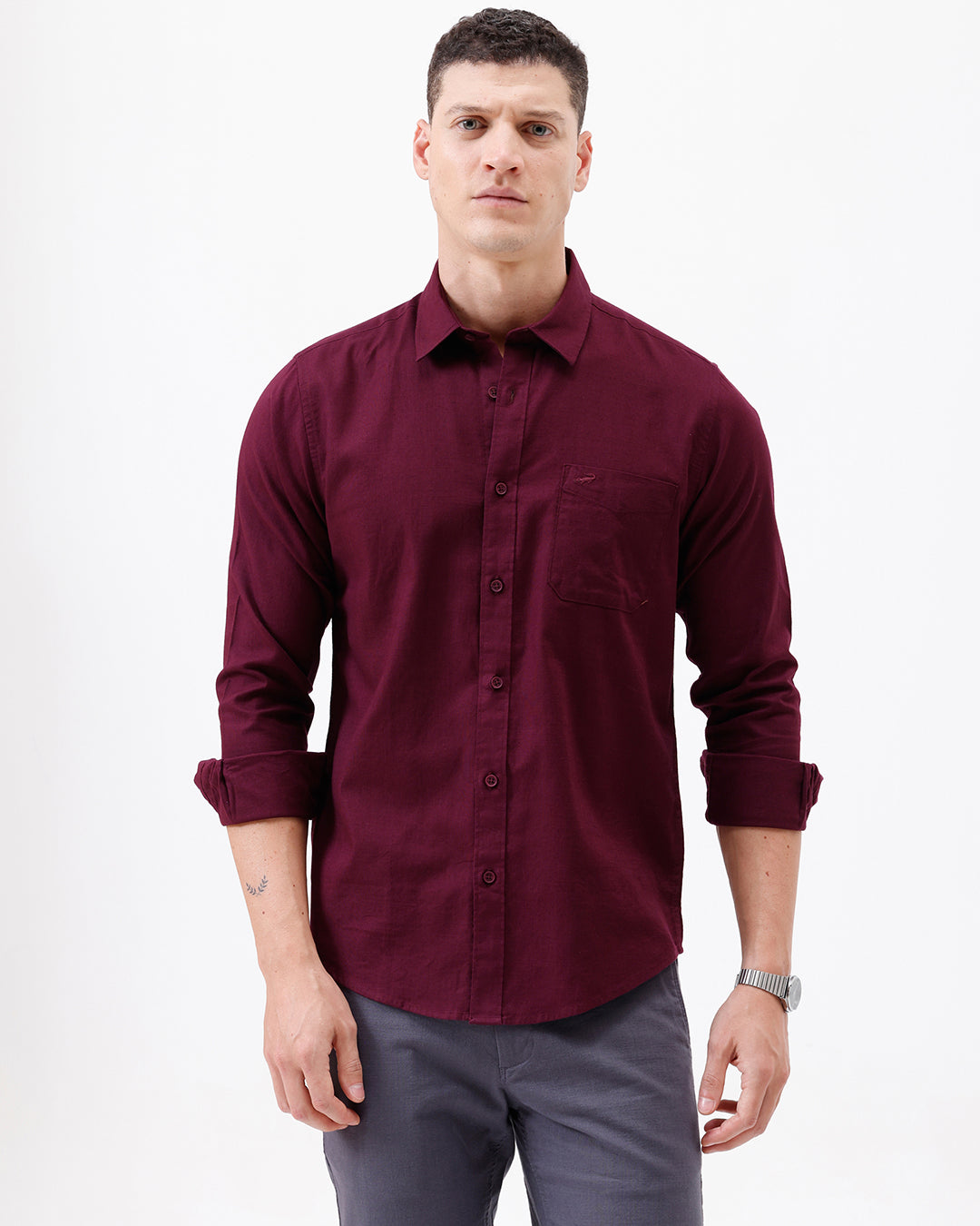 A man wearing a long-sleeve button-down shirt in deep burgundy with rolled-up sleeves and gray trousers, standing against a plain white studio background.