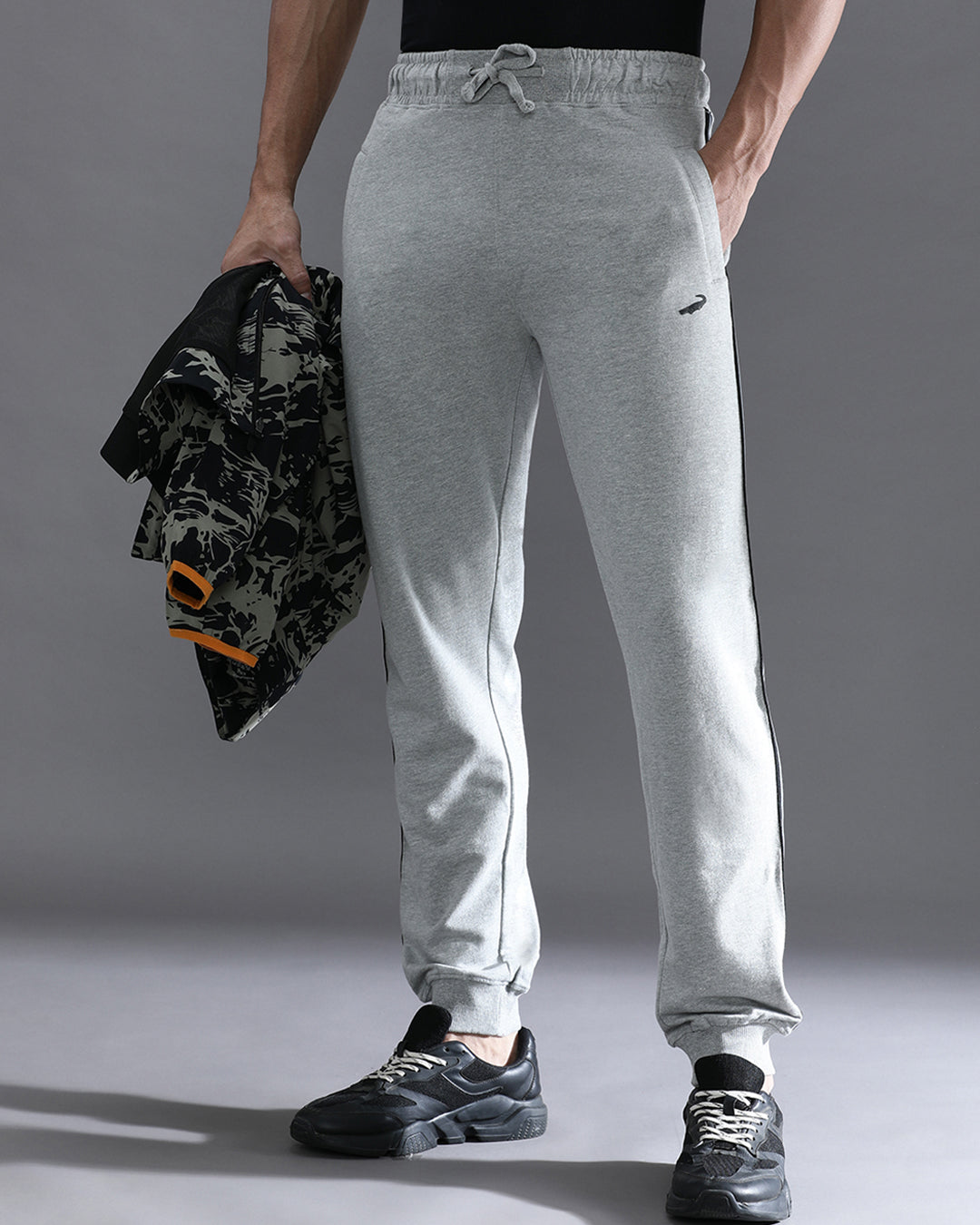 A person standing wearing grey melange solid classic fit joggers with a drawstring waist, holding a jacket and wearing sneakers.