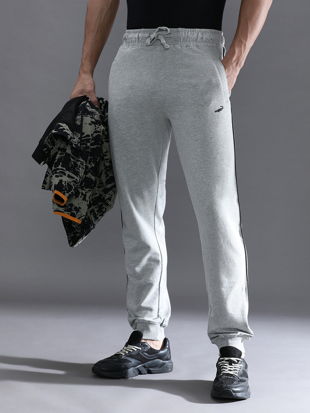 A person standing wearing grey melange solid classic fit joggers with a drawstring waist, holding a jacket and wearing sneakers.