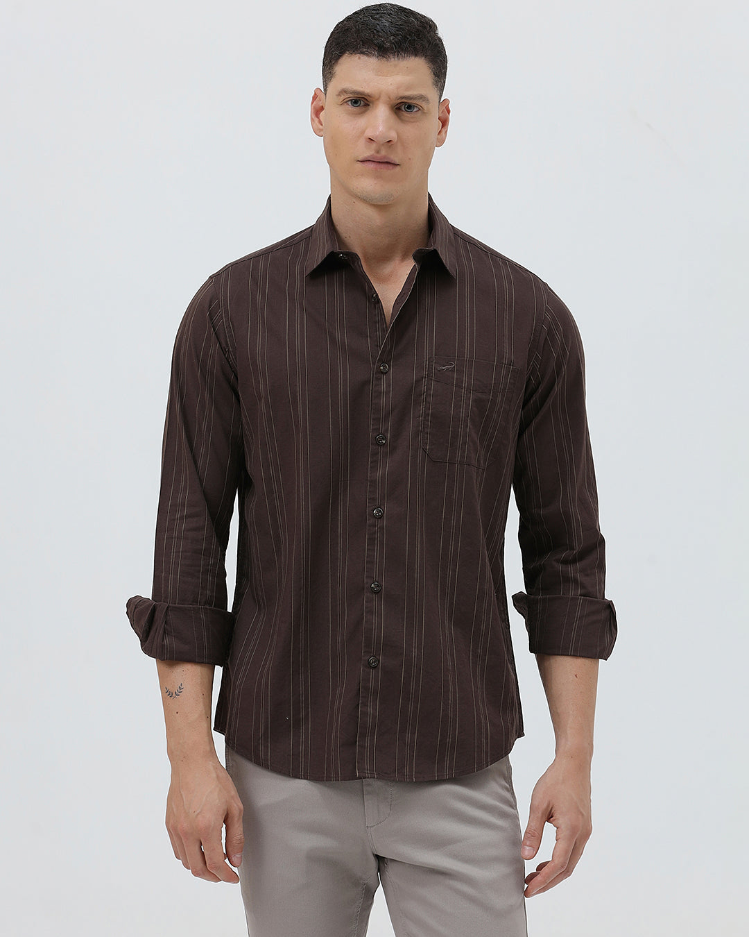 A man wearing a dark brown long-sleeve button-up shirt with thin vertical stripes and light gray trousers, standing against a plain light gray background.