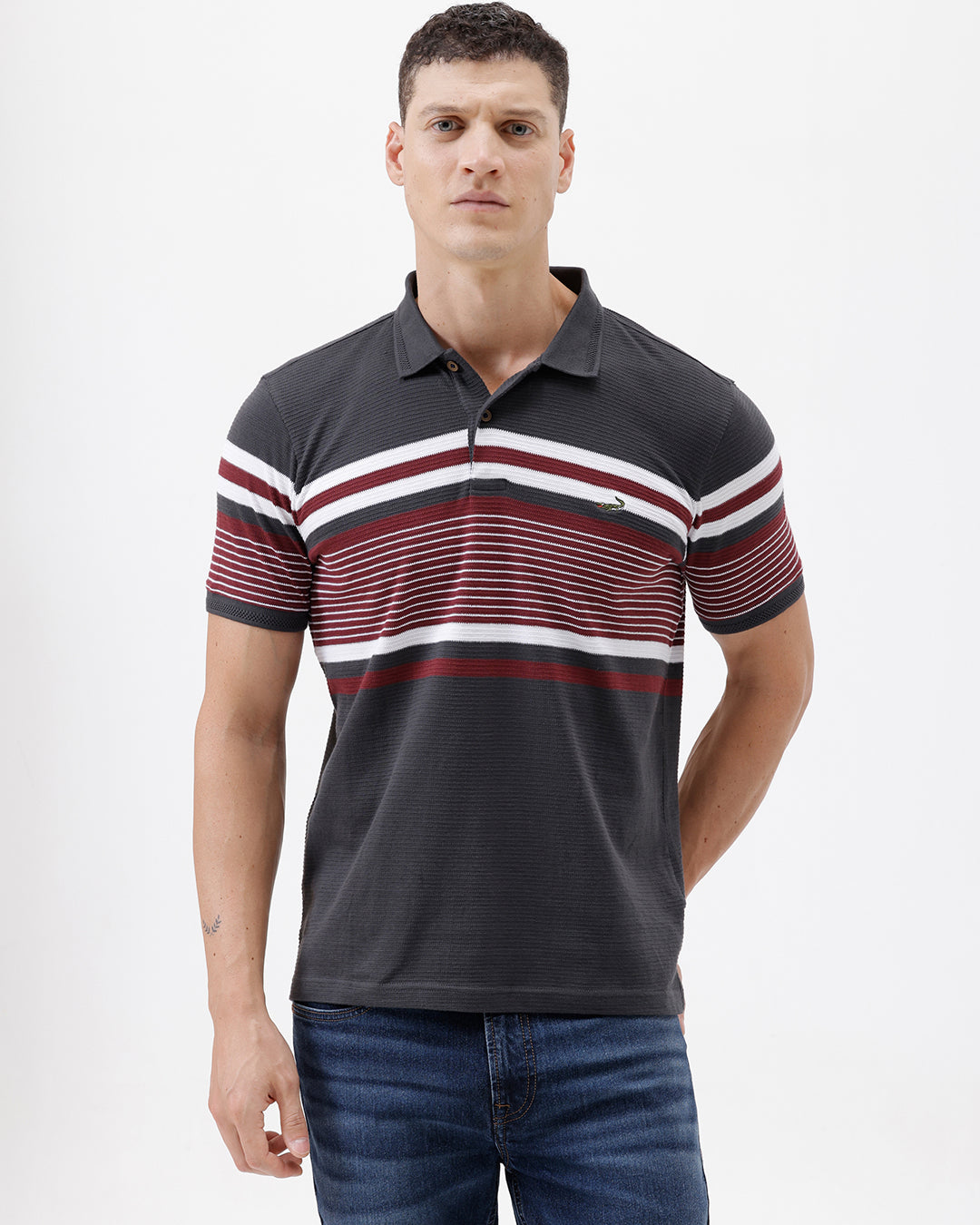 A man wearing a dark gray Lacoste polo shirt with horizontal red and white stripes across the chest and sleeves, paired with blue jeans, against a plain light gray background.