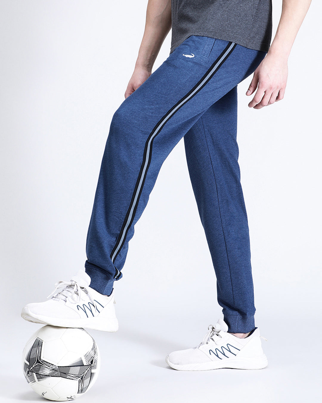 A person standing with one foot on a soccer ball, wearing blue melange solid classic fit joggers and white sneakers.