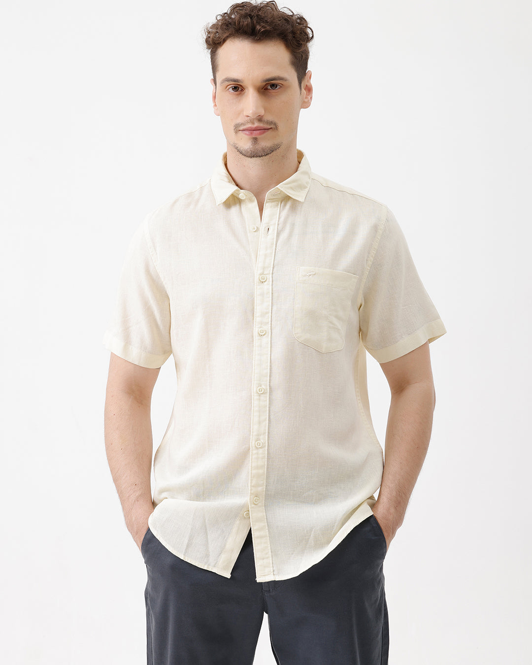 A man wearing a light cream-colored short-sleeve linen button-up shirt with a chest pocket, paired with dark trousers, against a plain white studio background.