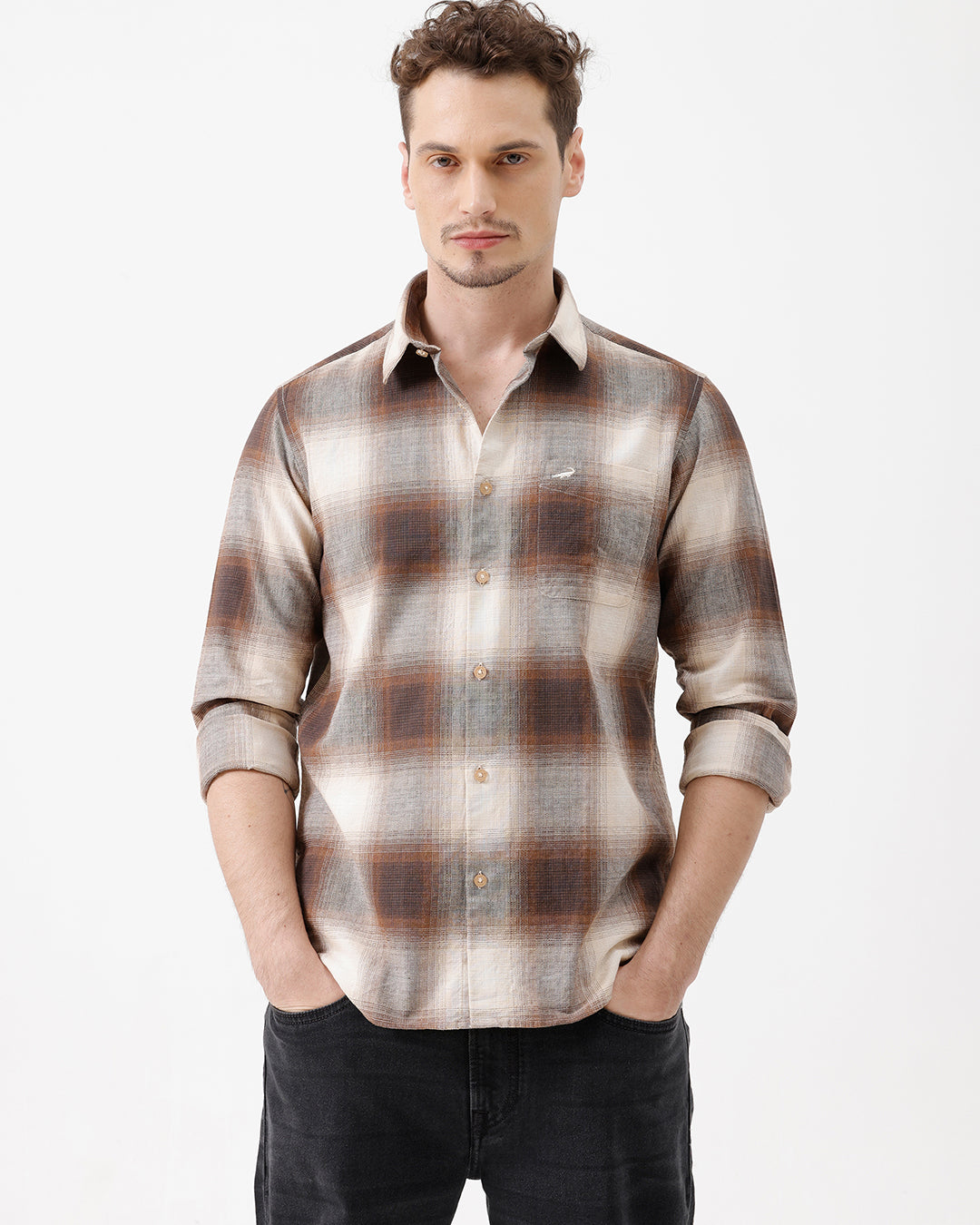 A man wearing a long-sleeve button-up plaid shirt in brown, beige, and gray tones, paired with dark jeans, standing against a plain white studio background.