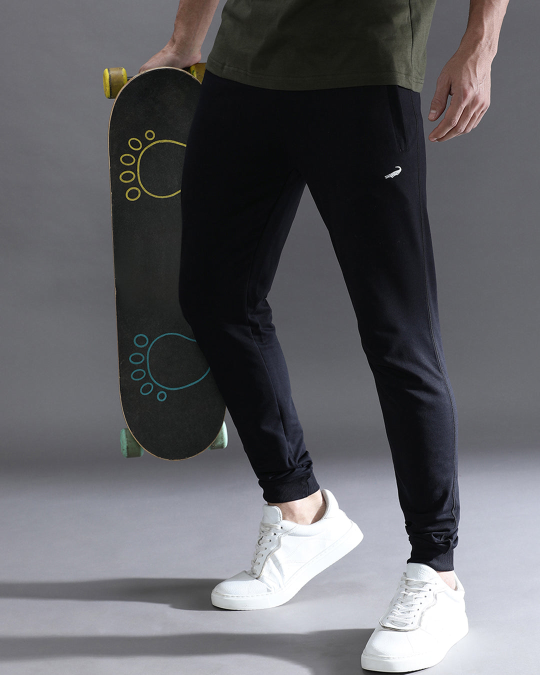 Person holding a skateboard wearing black pants with a Nike logo and white sneakers on a gray background