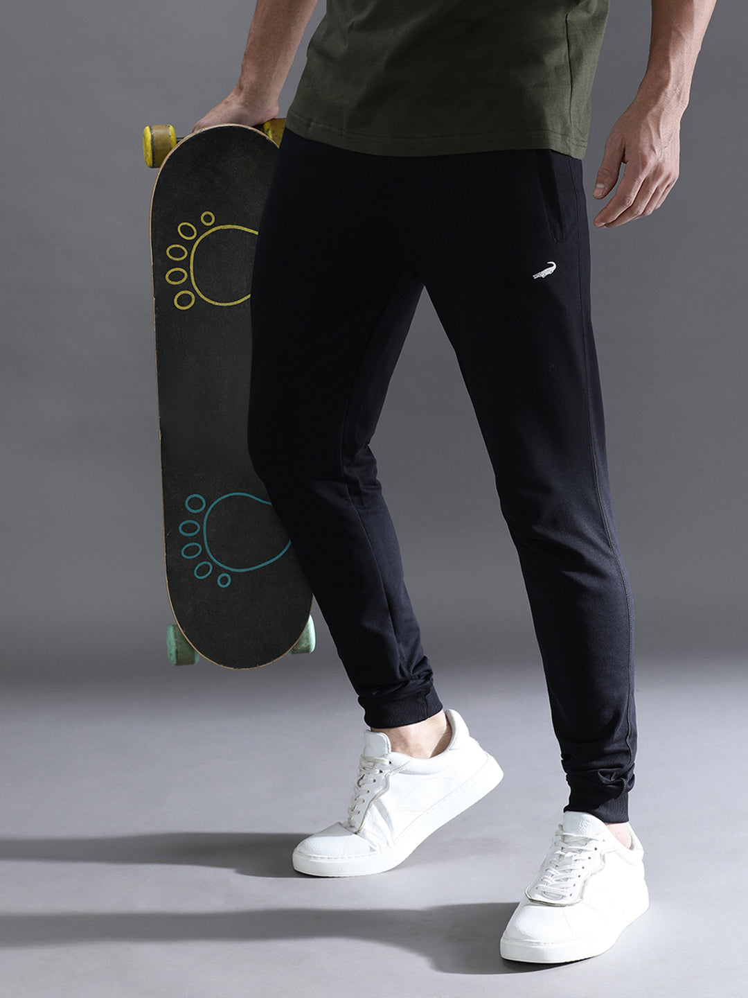 Person holding a skateboard wearing black pants with a Nike logo and white sneakers on a gray background