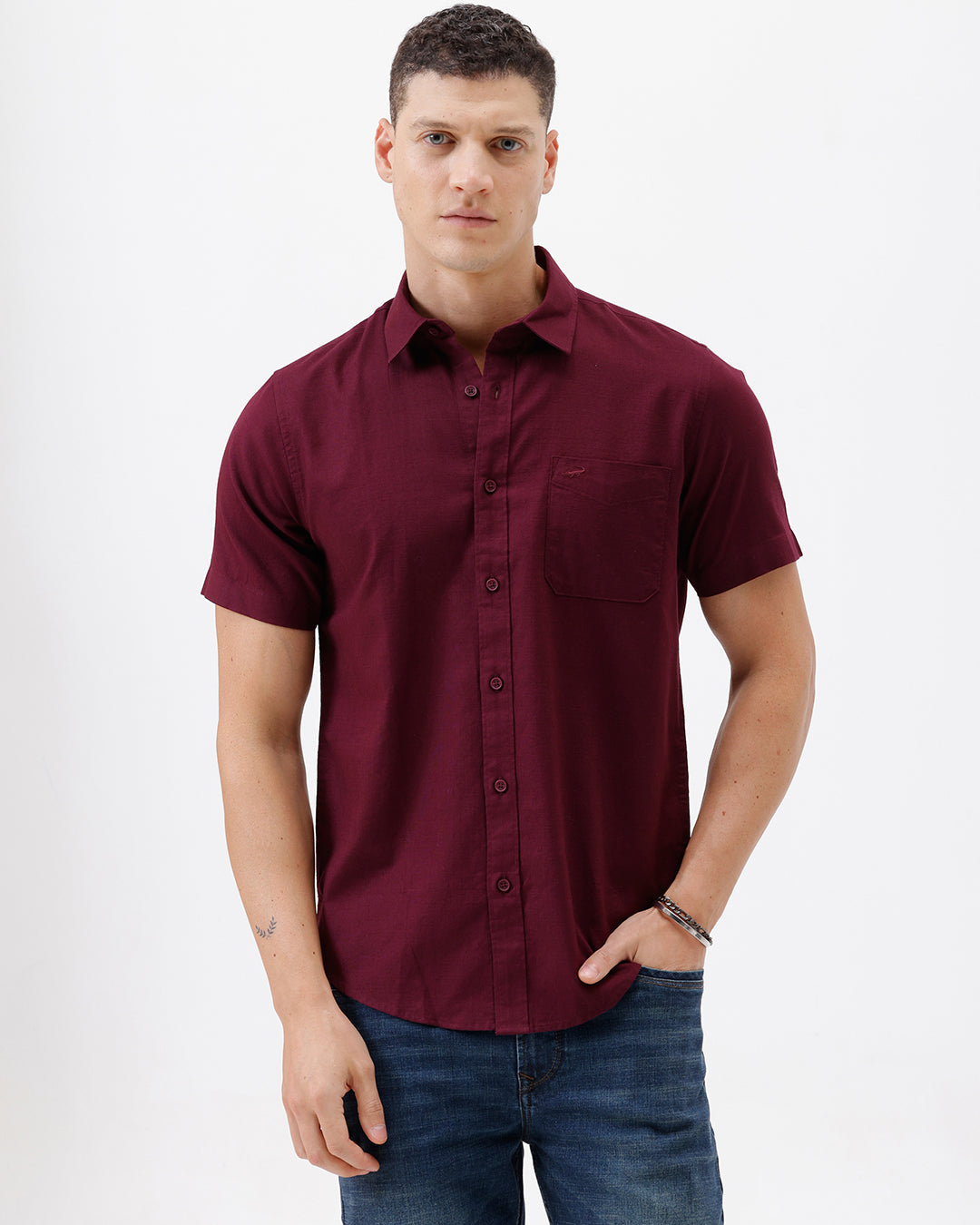 A man wearing a short-sleeve button-up shirt in maroon with a chest pocket, paired with blue jeans, standing against a plain white studio background.
