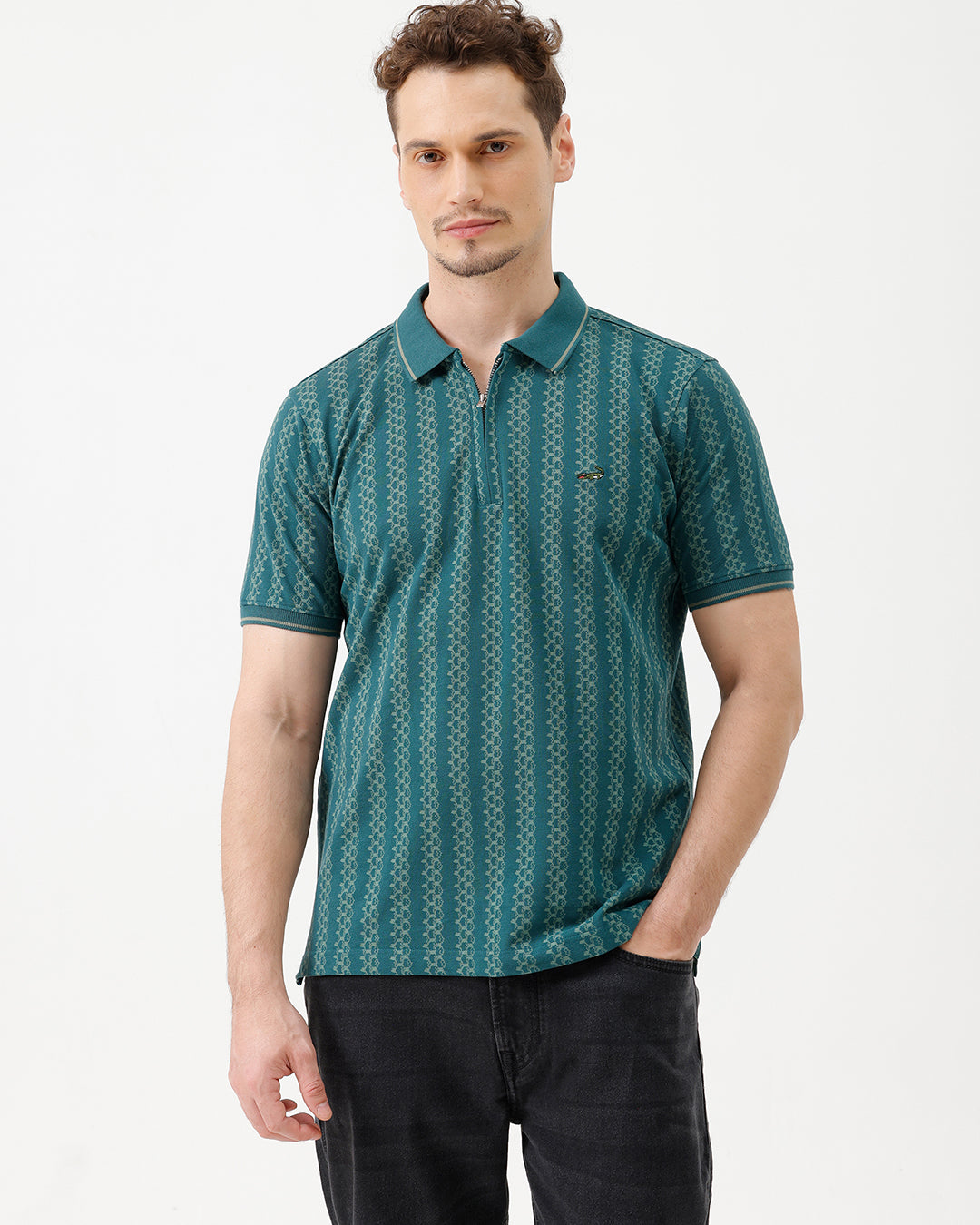 A man wearing a teal green short-sleeve polo shirt with a white geometric pattern, paired with black jeans, standing against a plain white studio background.