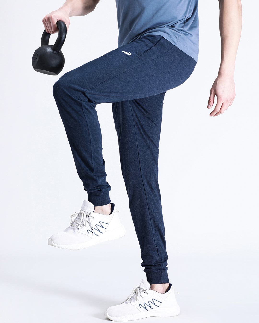 A person standing wearing navy blue joggers and white sneakers, holding a kettlebell in their left hand.
