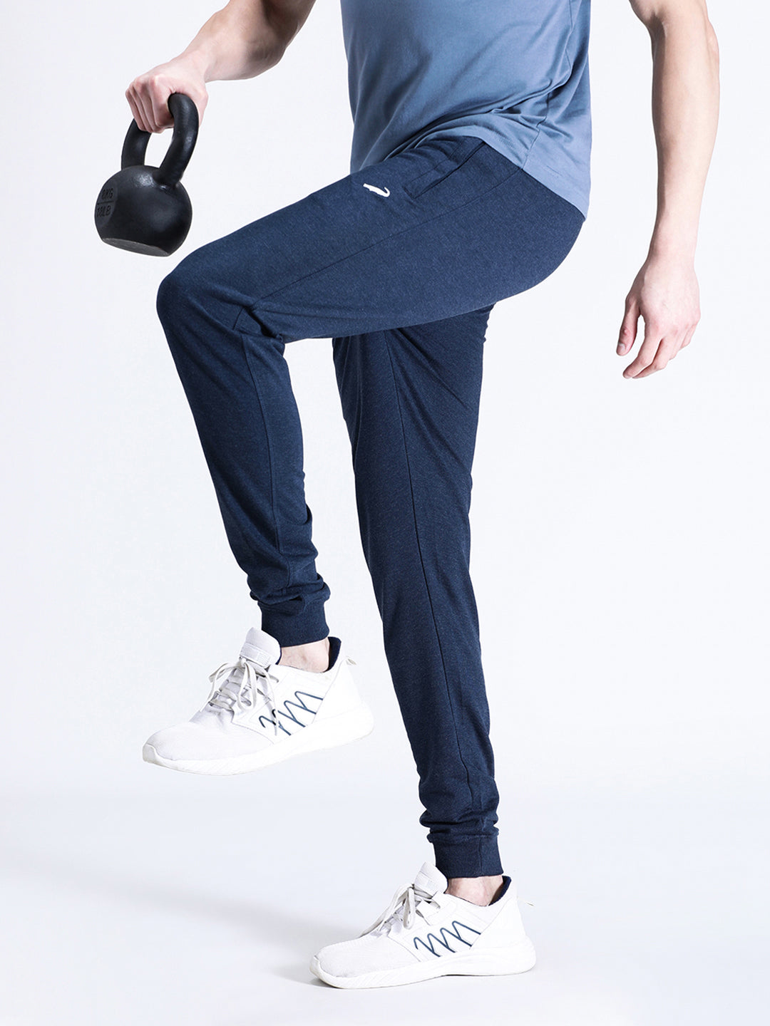 A person standing wearing navy blue joggers and white sneakers, holding a kettlebell in their left hand.