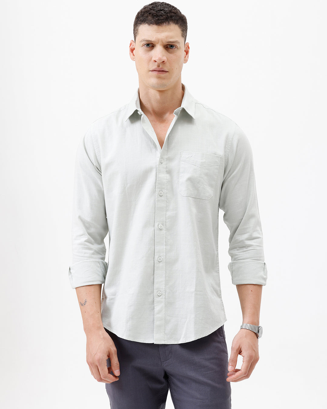 A man wearing a light sage green long-sleeve button-down shirt with rolled-up sleeves and dark gray trousers, standing against a plain white studio background.