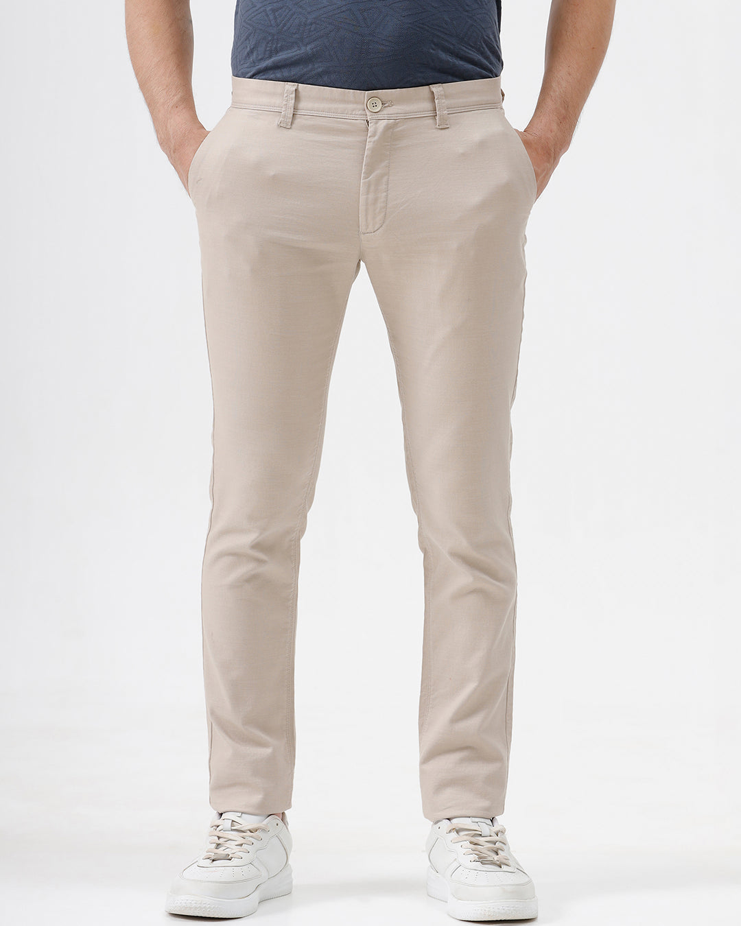A person wearing slim-fit beige chinos with a blue patterned t-shirt and white sneakers, standing against a white studio background.