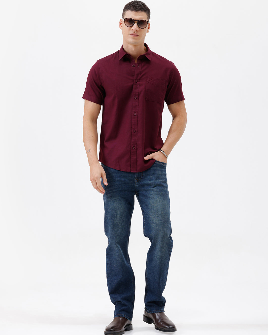 A man wearing a short-sleeve button-up shirt in maroon with a chest pocket, paired with blue jeans, standing against a plain white studio background.