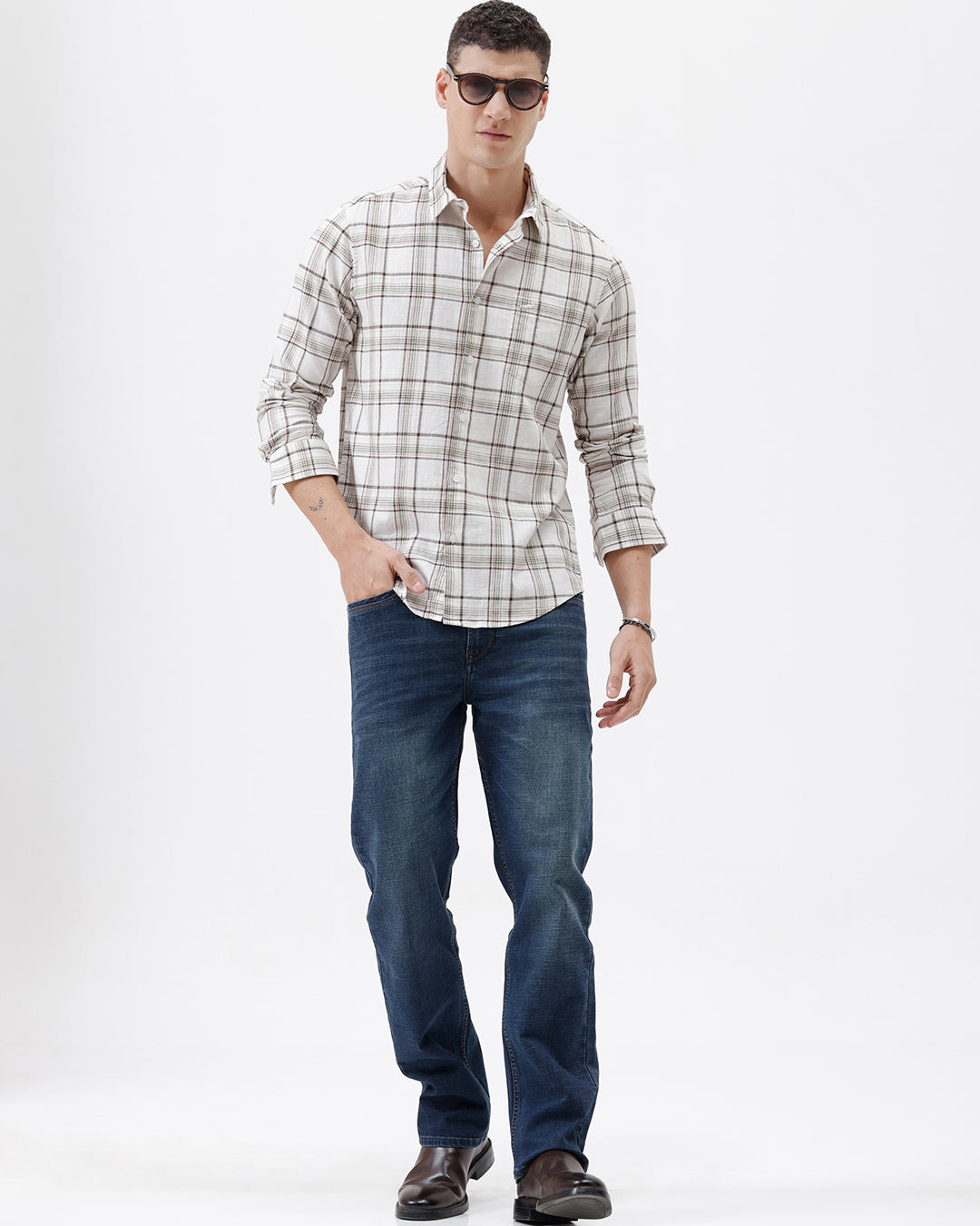 A man wearing a long-sleeve plaid button-up shirt in beige and brown with dark blue jeans, standing against a plain white studio background.