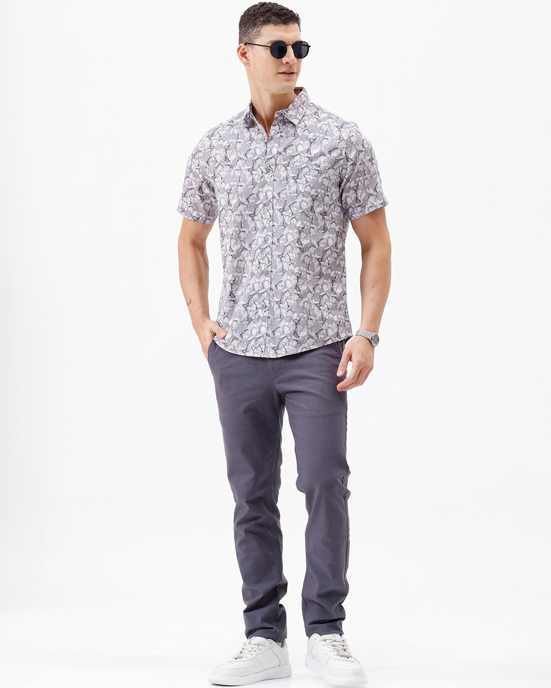 A man wearing a short-sleeve button-up shirt with a gray and white abstract pattern, paired with dark gray trousers, standing against a plain white studio background.