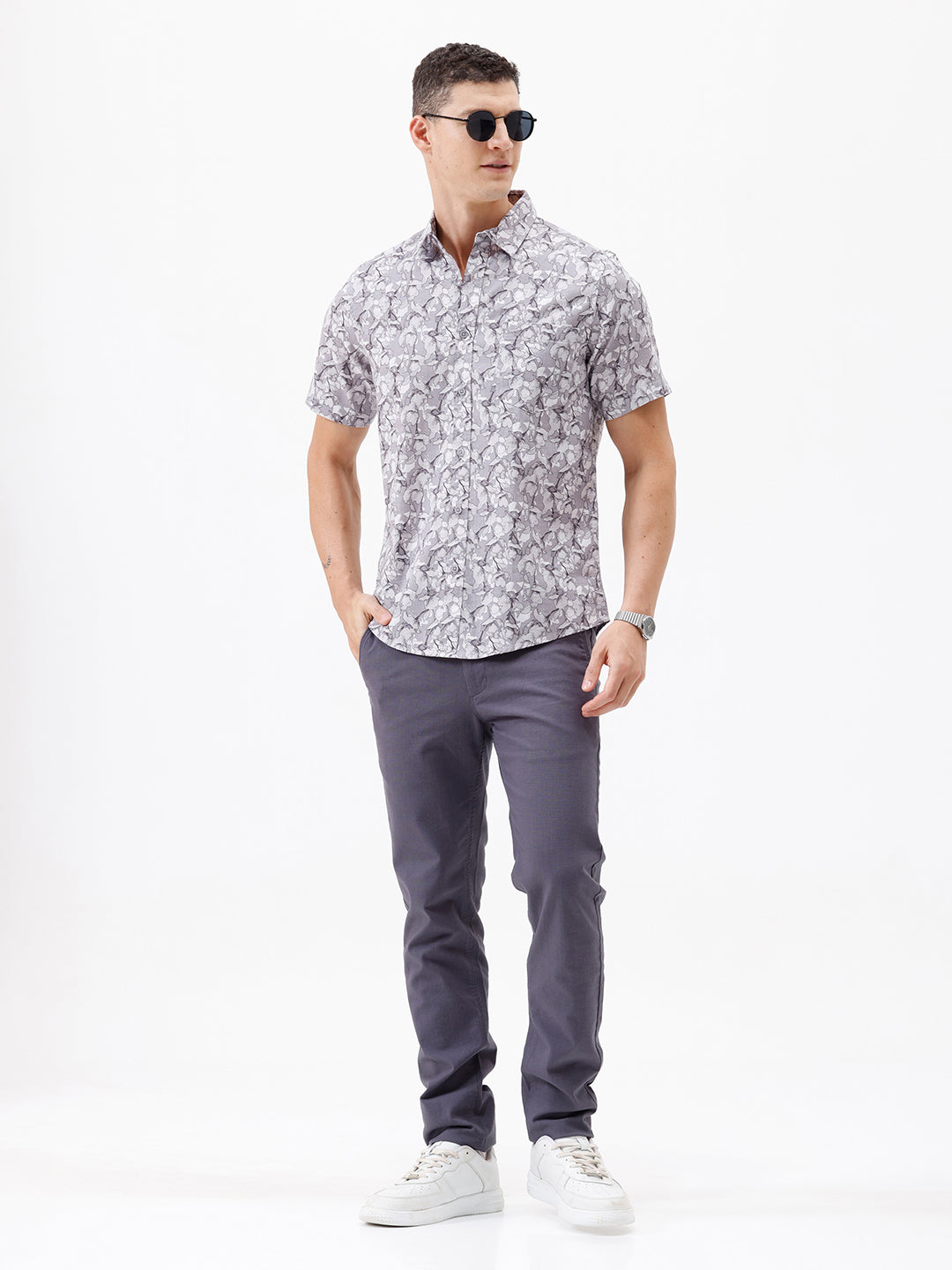 A man wearing a short-sleeve button-up shirt with a gray and white abstract pattern, paired with dark gray trousers, standing against a plain white studio background.