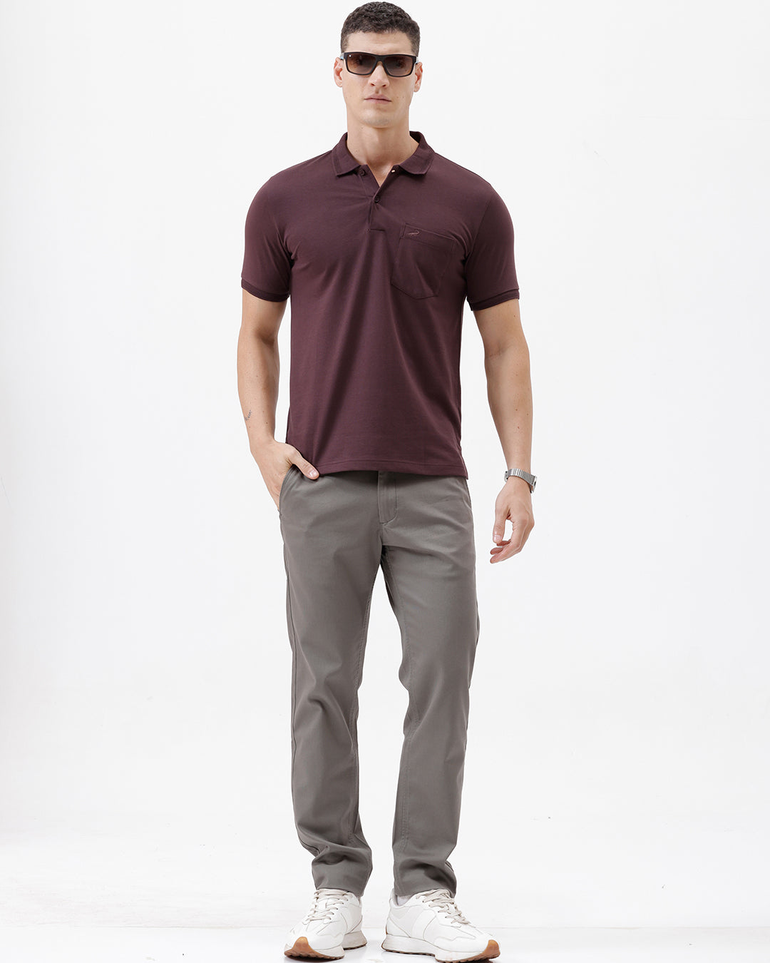 A man wearing a short-sleeve polo shirt in dark burgundy with a chest pocket, paired with gray trousers, standing against a plain light gray background.